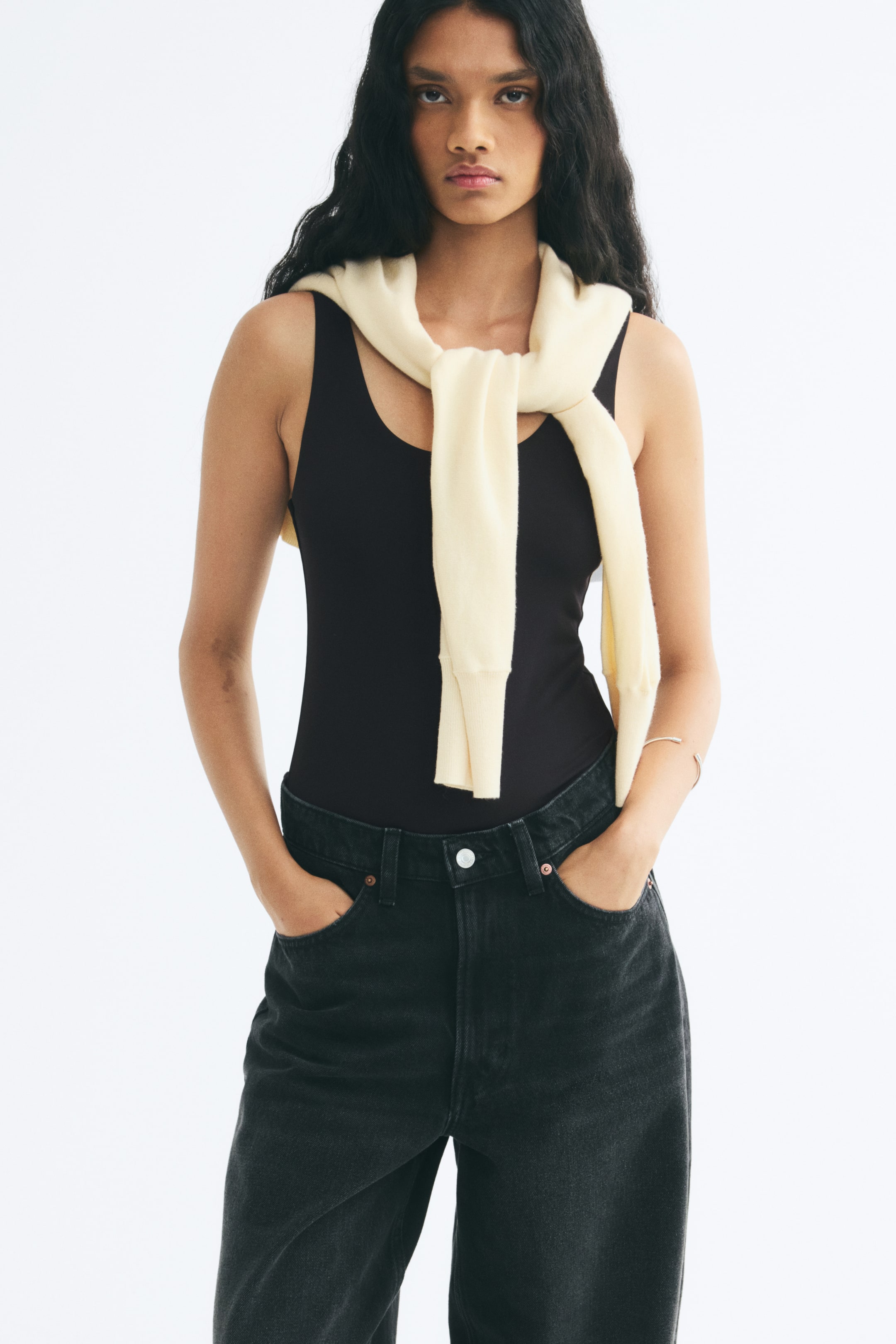View larger image: A black stretch jersey bodysuit with a scoop neckline and wide shoulder straps is worn by the model. The bodysuit is tucked into black jeans and styled with a cream-colored sweater draped over the shoulders.