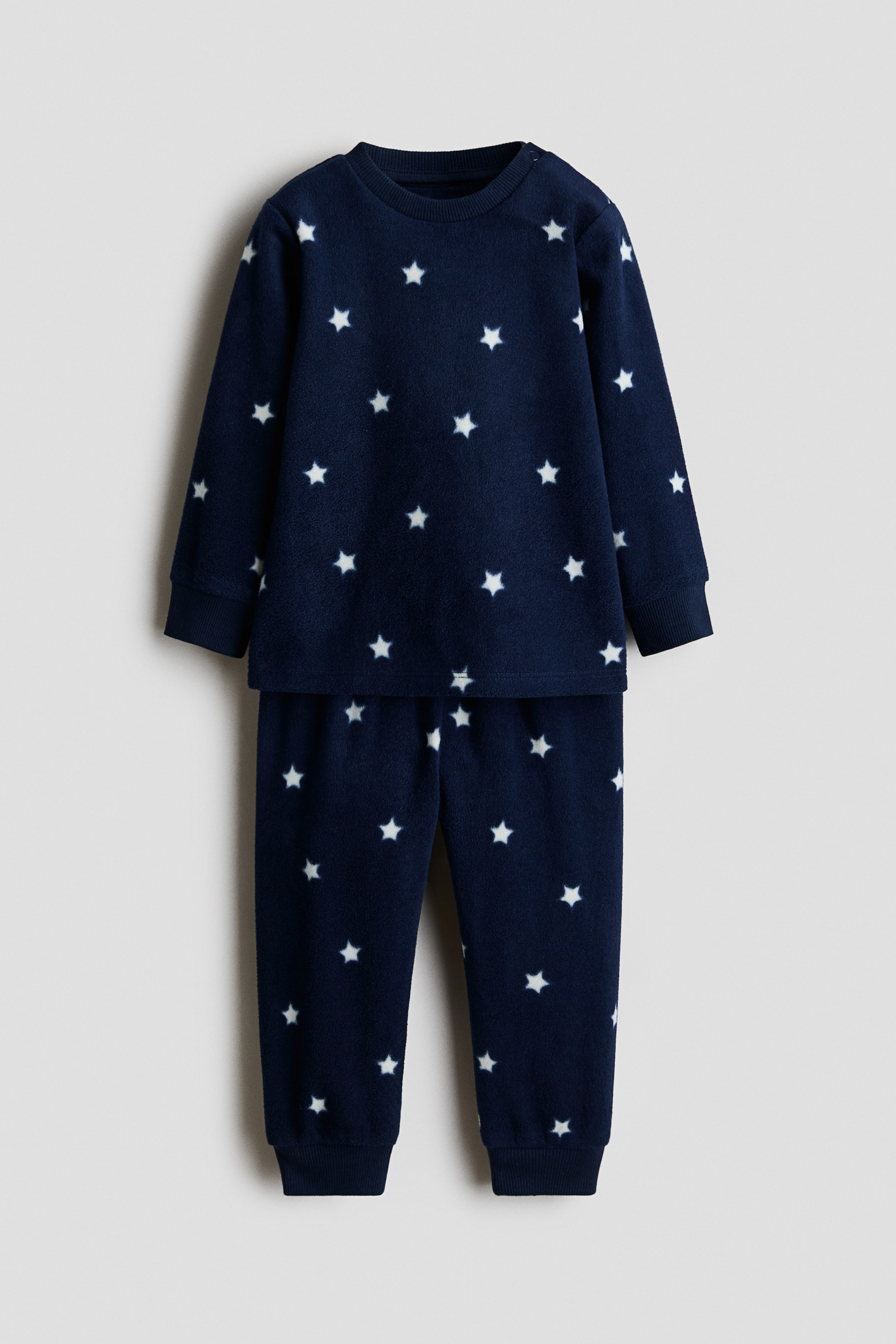 View larger image: A navy blue fleece two-piece pyjama set is shown front-facing, featuring a repeating pattern of white stars. The long-sleeved top has a crew neck, ribbed cuffs, and a ribbed hem, while the long pants are designed with ribbed cuffs at the ankles.