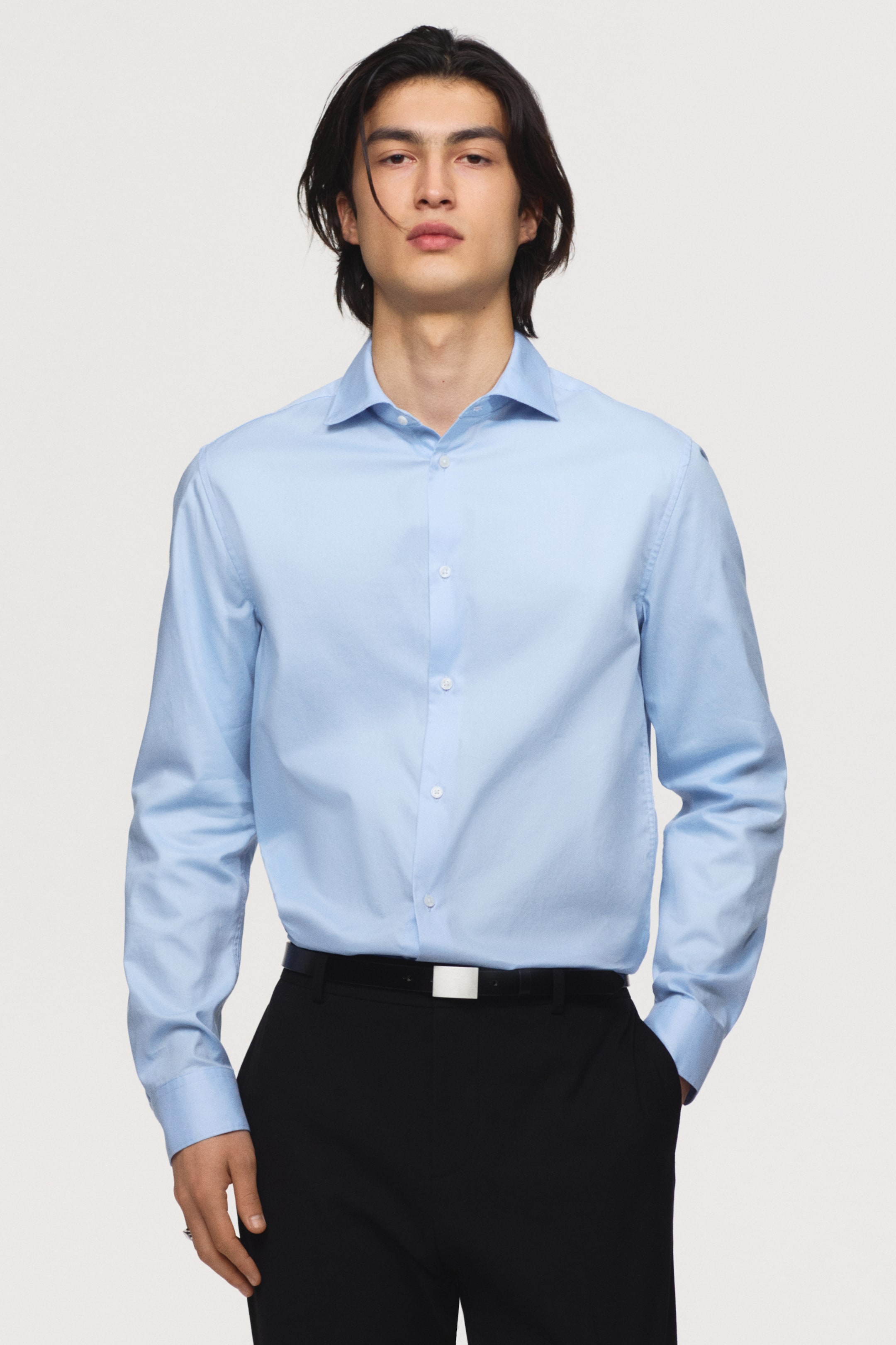 View larger image: A light blue slim-fit shirt with a cut-away collar and French front is being worn by the model. The long-sleeved shirt features buttoned cuffs and is tucked into black trousers, with the model standing with one hand in a trouser pocket.