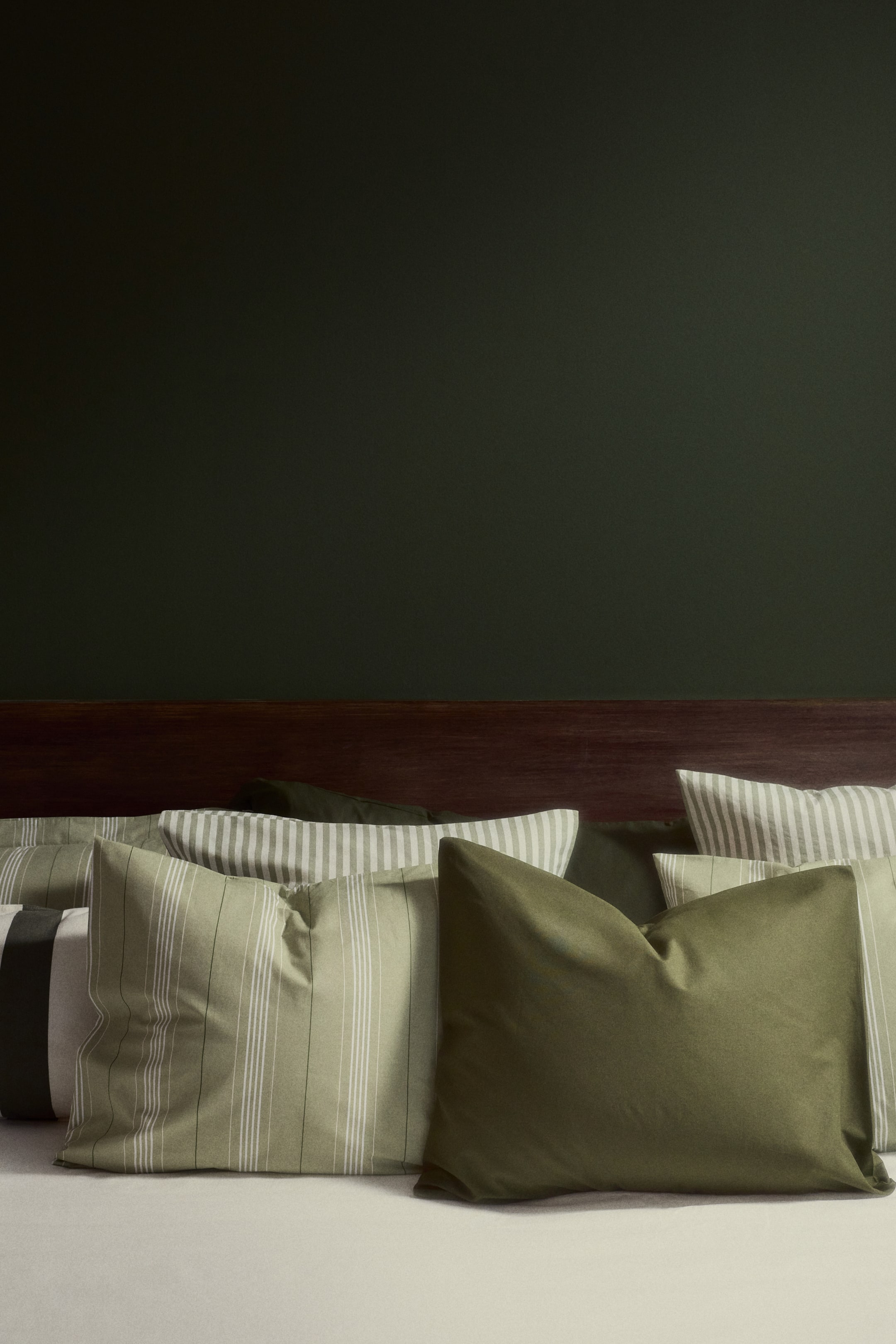 View larger image: Multiple pillows are arranged in a layered display, facing forward. Some pillows appear in solid olive green, while others feature thin white vertical stripes or wider light gray horizontal stripes on light green fabric, with a black-and-white striped pillow visible at the far left.