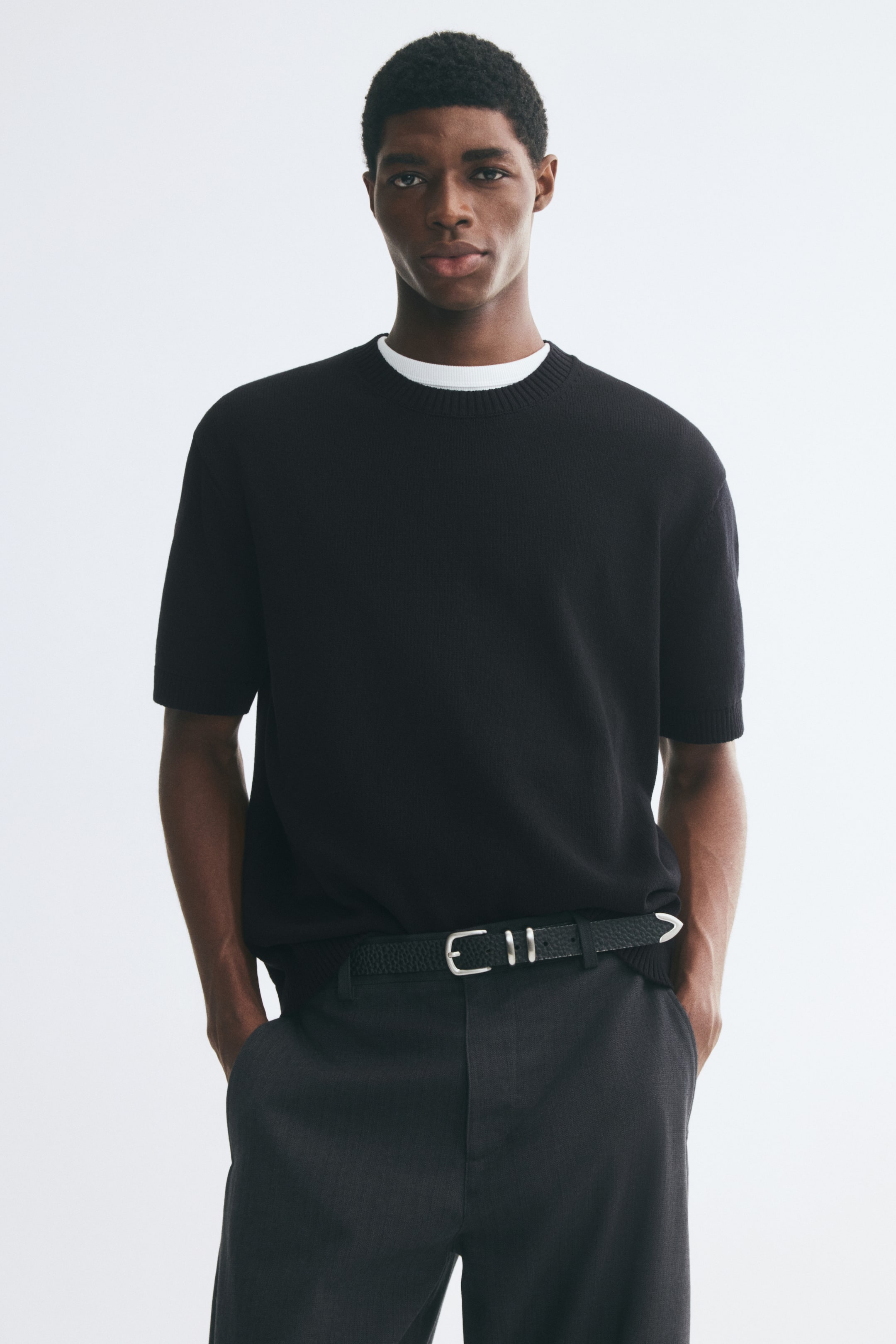 View larger image: The model is wearing a black short-sleeve knit tee layered over a visible white crew-neck T-shirt. The tee features a ribbed crew neckline and ribbed cuffs, and is styled with dark gray trousers and a black belt.