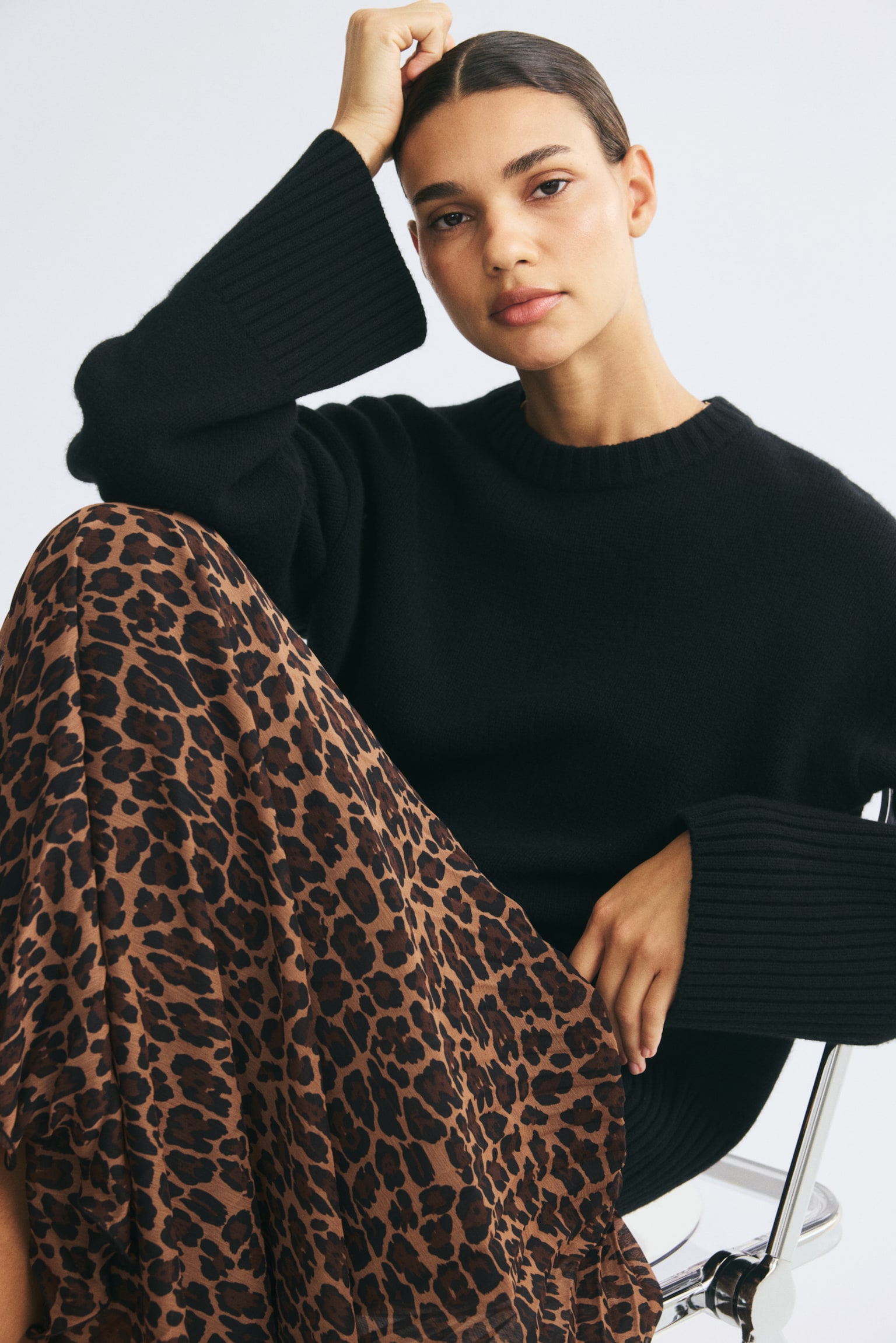 A leopard-print, pleated fabric skirt is being worn by the model while sitting. The loose-fitting skirt is styled with a long-sleeved black knitted sweater. - 1
