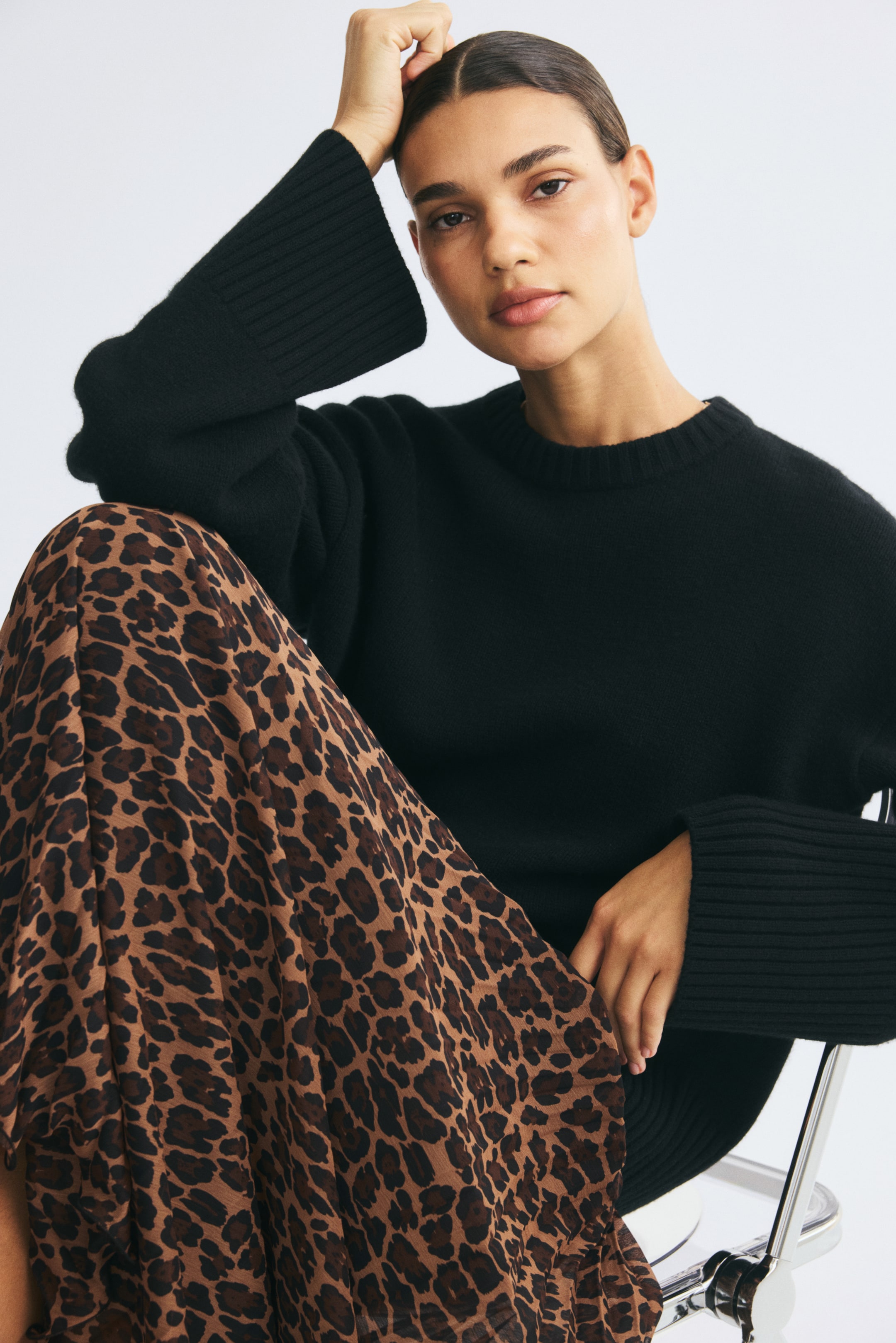 Größeres Bild ansehen: Ein Rock aus plissiertem Stoff mit Leopardenmuster wird von dem Model im Sitzen getragen. Der locker sitzende Rock ist mit einem schwarzen Strickpullover mit langen Ärmeln kombiniert.