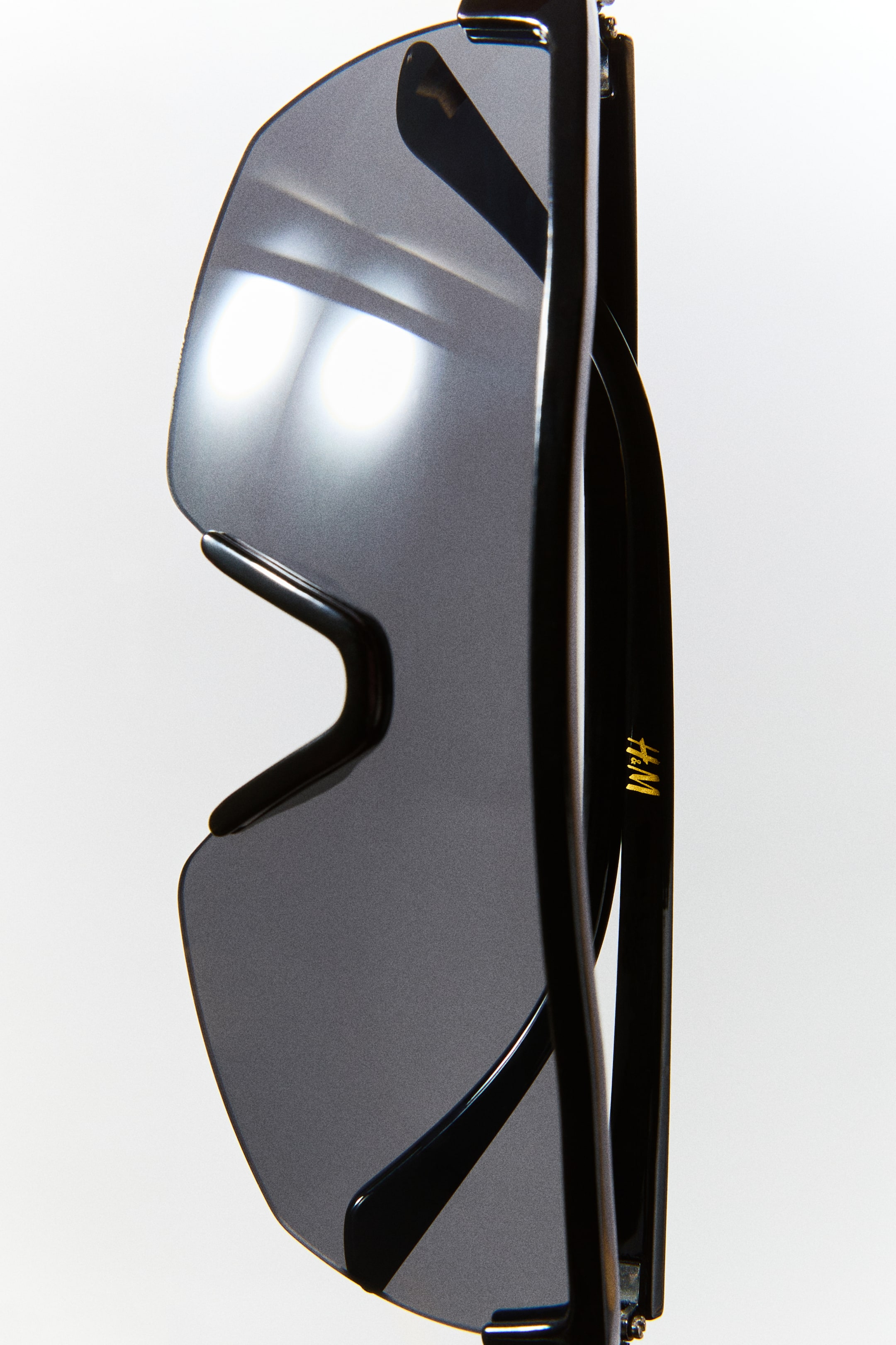 View larger image: Shield sunglasses featuring a large, curved, gray-tinted plastic lens and a glossy black plastic frame present a wraparound design. The folded temples display a small gold logo on the side.