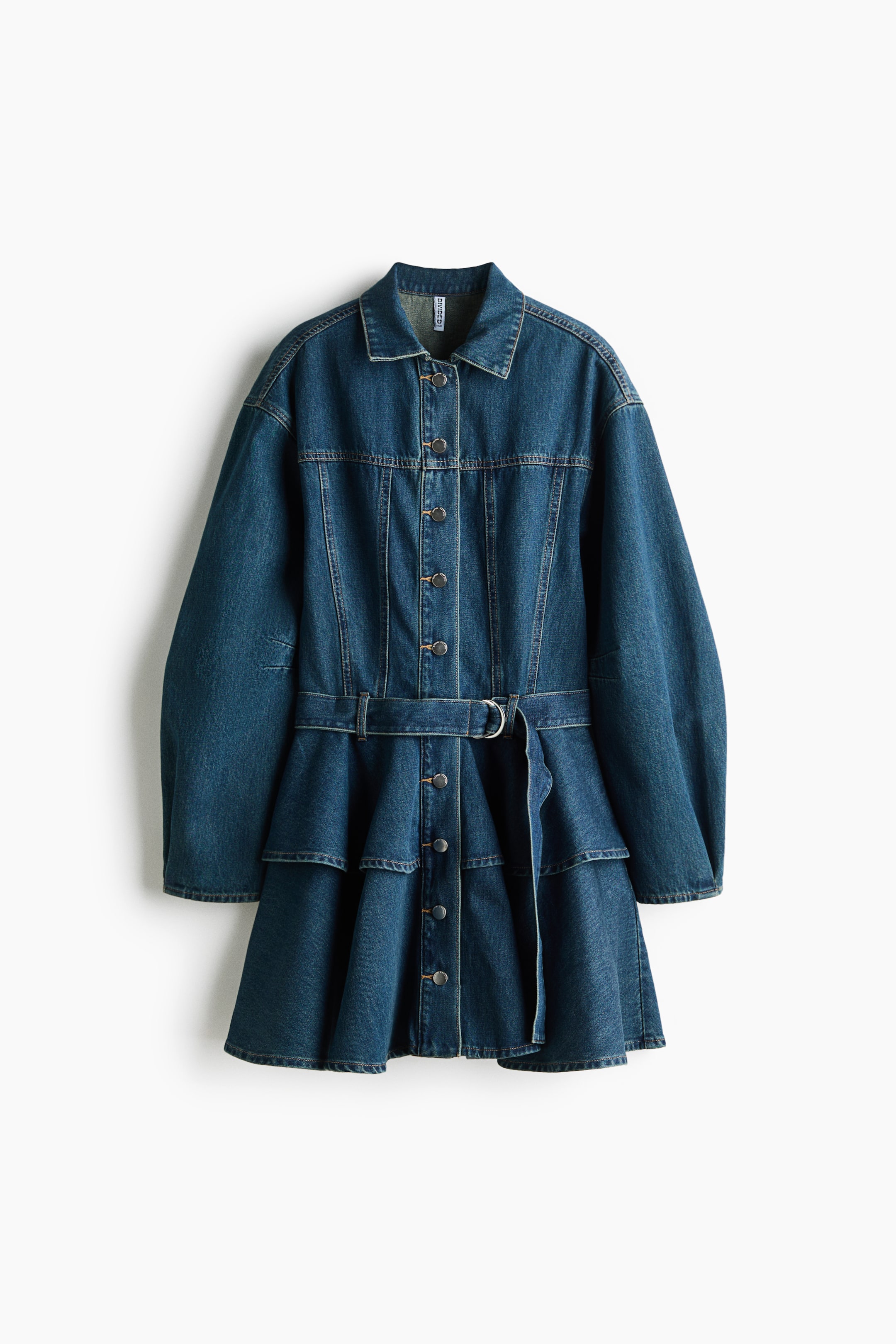 Vezi imaginea mai mare: O rochie din denim albastru închis, văzută din față, prezintă un decolteu cu guler, o închidere completă cu nasturi în partea din față, cu nasturi de culoare deschisă, și o cusătură vizibilă de tip yoke pe piept. Este concepută cu mâneci lungi voluminoase și este strânsă în talie cu o curea asortată din denim, prevăzută cu o cataramă D-ring în nuanță argintie. Fusta are două volane dispuse în trepte.
