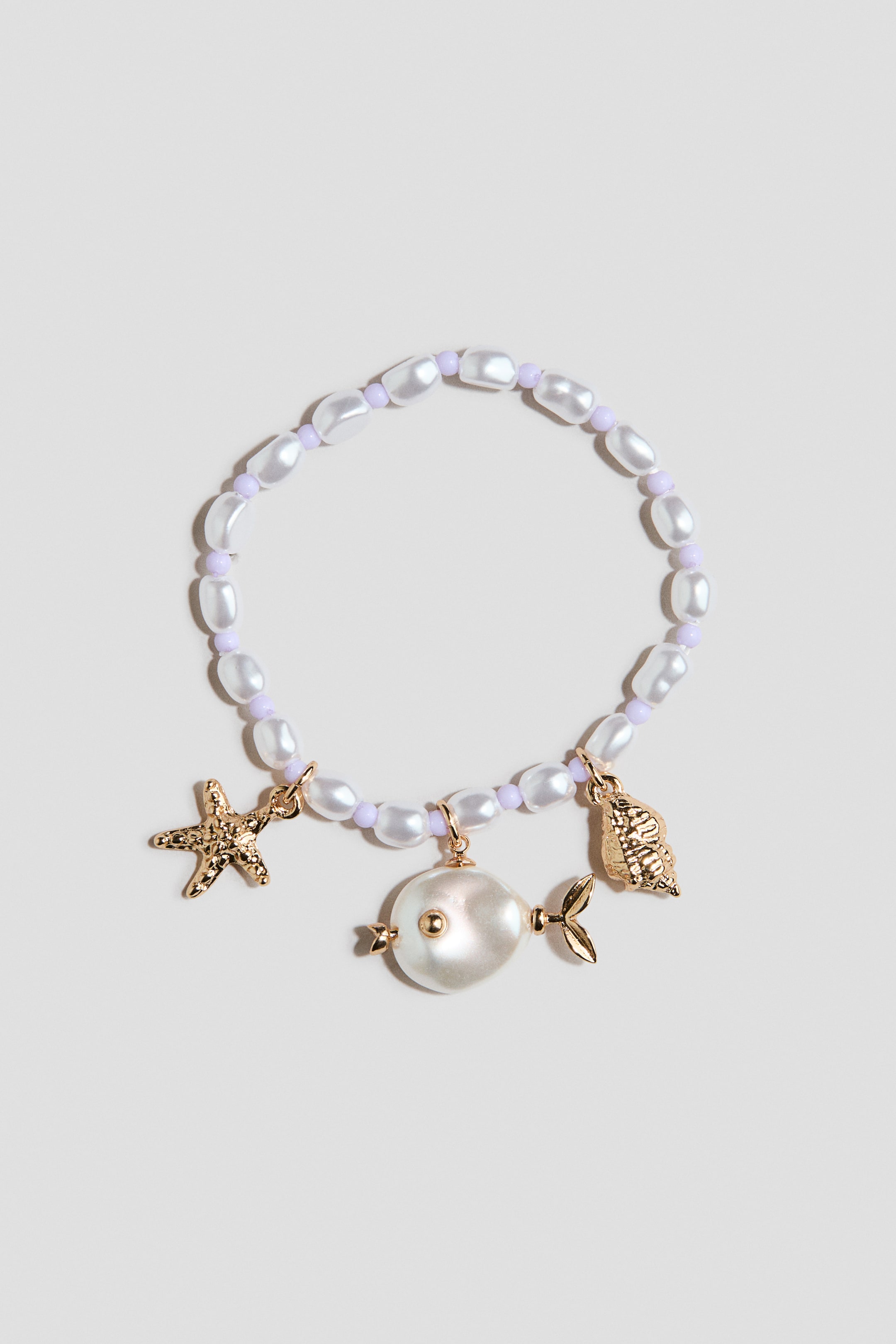 Größeres Bild ansehen: Das Armband präsentiert weiße, unregelmäßig geformte Perlen, die mit kleinen violetten Perlen durchsetzt sind, und wird von vorne gezeigt. Drei goldfarbene Anhänger baumeln daran, darunter ein strukturierter Seestern, eine detaillierte Muschel und ein Fischanhänger mit einem großen, flachen Perlenkörper und goldfarbenen Flossen.