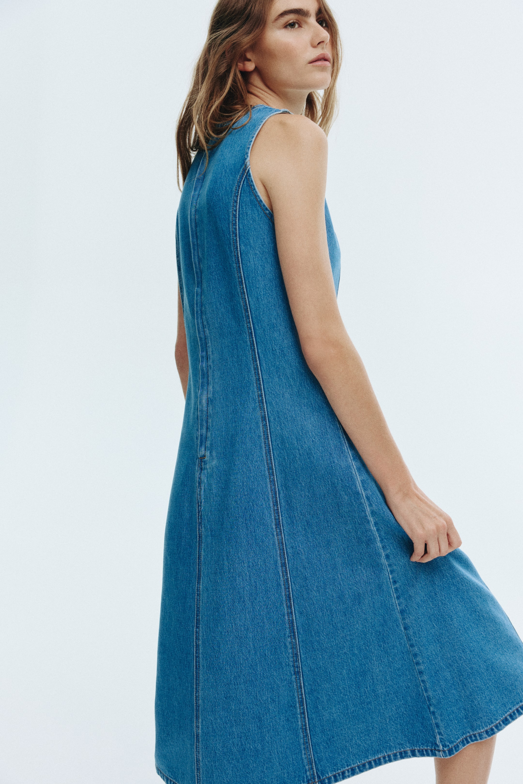 View larger image: A medium blue denim A-line dress is being viewed from a side-back angle, showcasing its sleeveless design. Prominent vertical seam lines are running down the back, highlighting its paneled construction and flared silhouette.