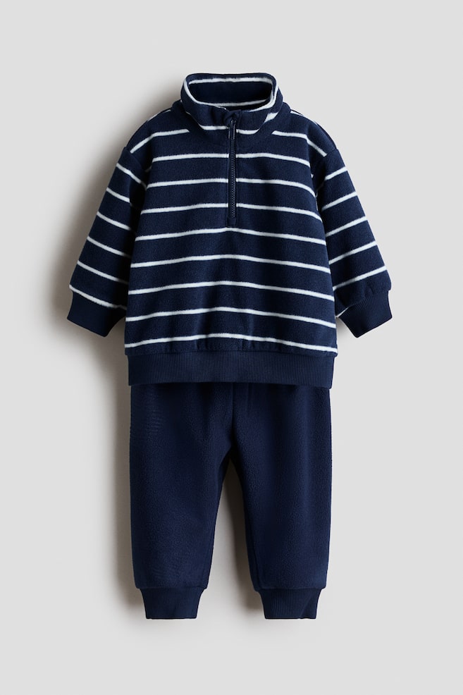 Ein zweiteiliges Fleece-Set in Frontansicht mit einem marineblauen Pullover mit weiÃen horizontalen Streifen, Stehkragen, Half-Zip-Verschluss, überschnittenen Schultern sowie gerippten Bündchen und Saum. Die dazu passenden einfarbig marineblauen Hosen vervollständigen das Set und verfügen über gerippte Knöchelbündchen. - 1