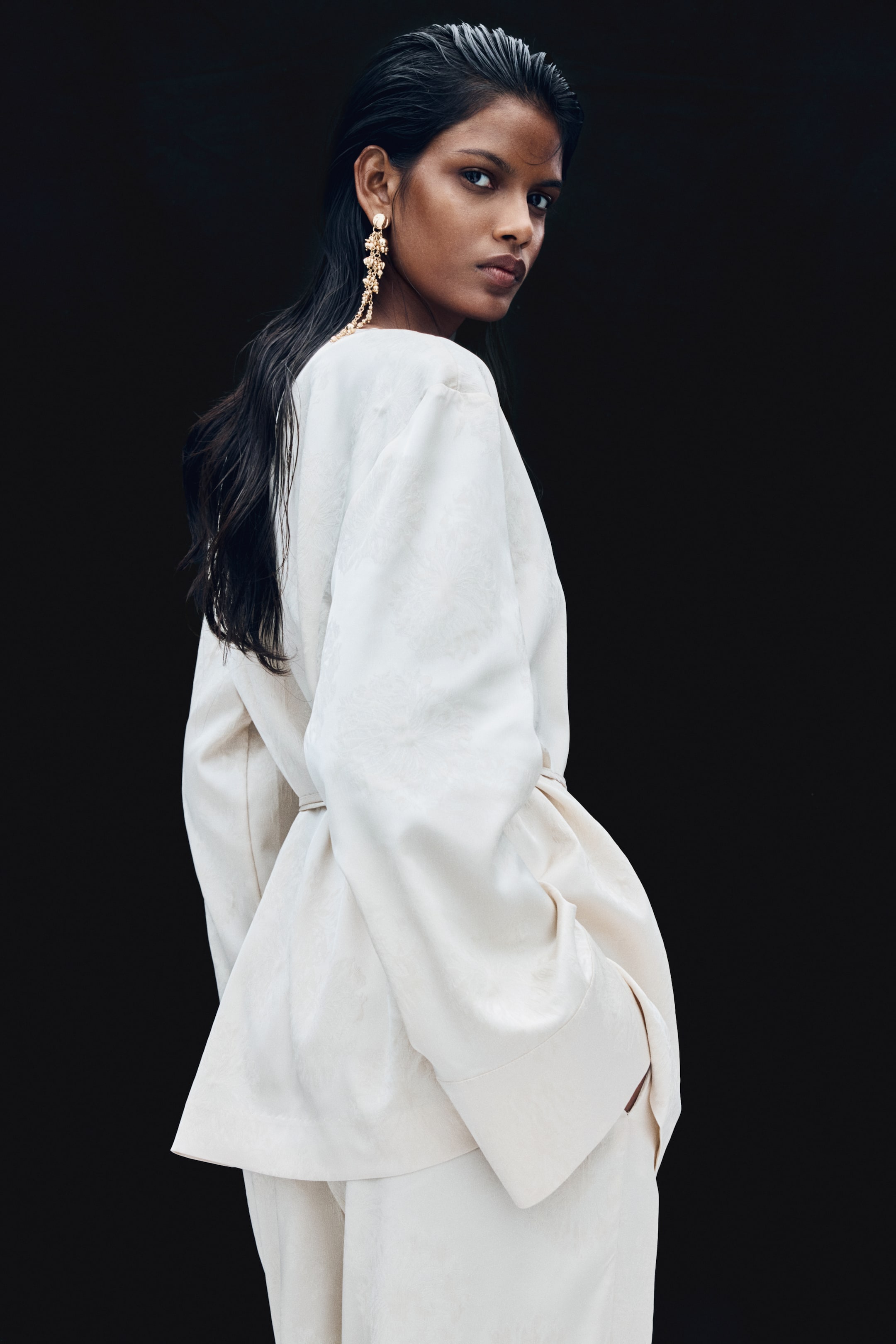 View larger image: An off-white, relaxed-fit blazer in a drapey jacquard weave with a slight sheen is being worn by the model. The blazer features long sleeves with wide, double-layered cuffs and a tied waist, and is paired with matching trousers.
