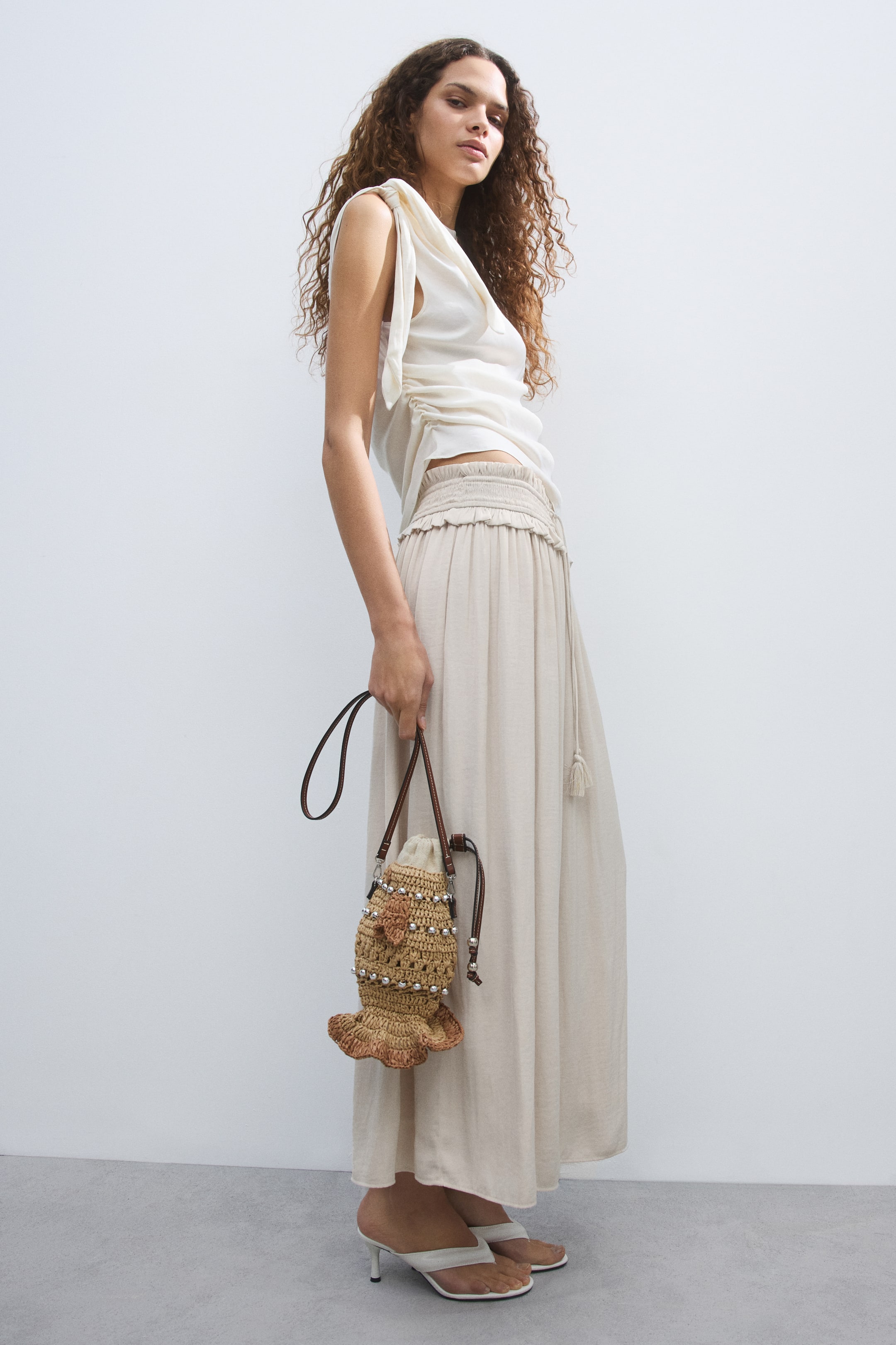 View larger image: The small bucket bag is being held by a hand, facing forward and slightly to the side. It is crafted from woven natural-toned material, featuring rows of silver-toned beads, a crocheted flower appliqué, and a ruffled bottom edge, with a thin dark brown strap.