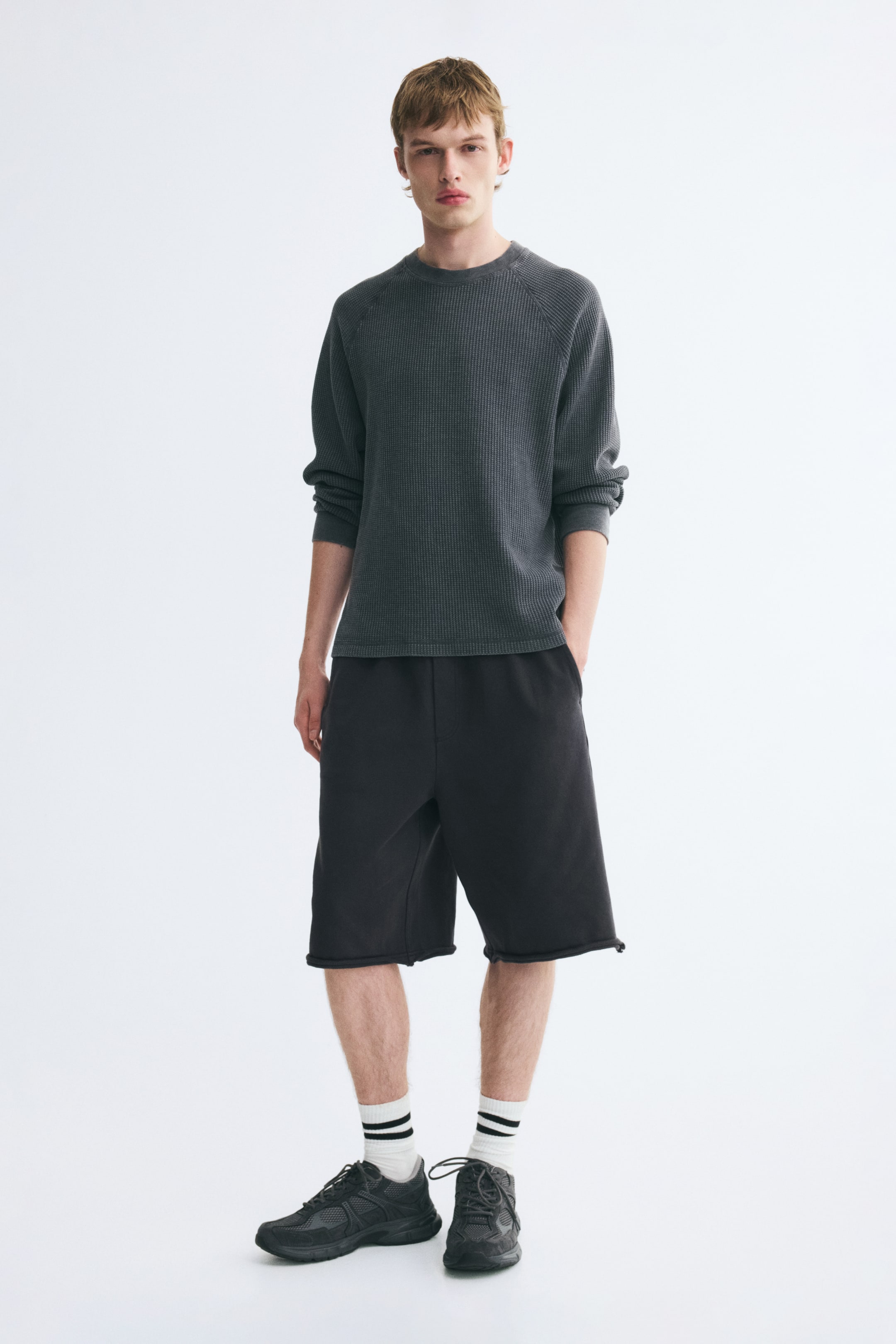 View larger image: A dark grey, waffle-knit long-sleeve crewneck top with raglan sleeves is being worn by the model. The top is styled with loose-fitting black shorts, white socks with black stripes, and black sneakers.