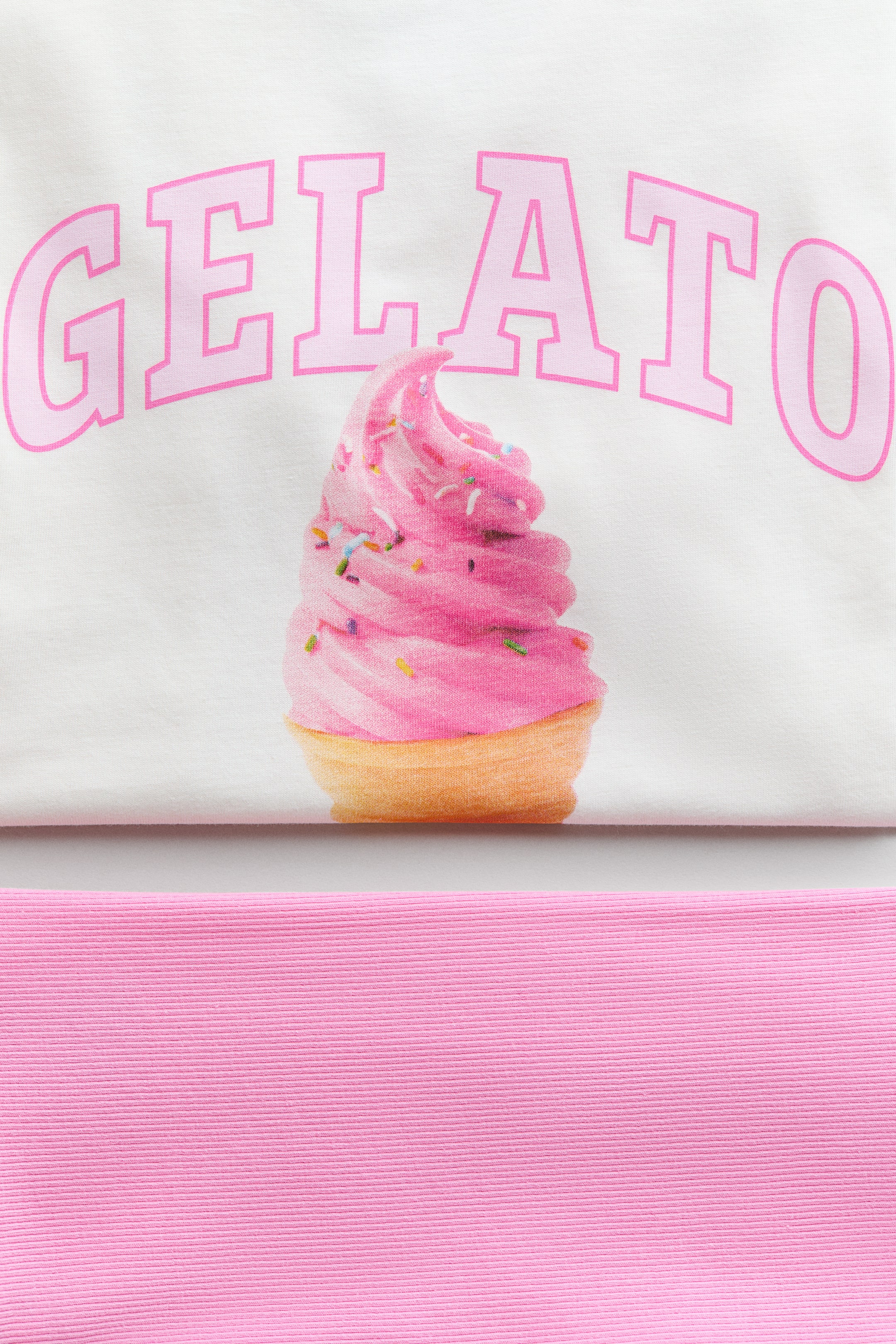 Visualizza l'immagine ingrandita: Un set che mostra una T-shirt bianca con una stampa ad arco della scritta "GELATO" in rosa sopra un ricciolo di gelato rosa con granelli colorati in un cono, posizionato sopra una sezione di pantaloncini da ciclismo rosa a coste.