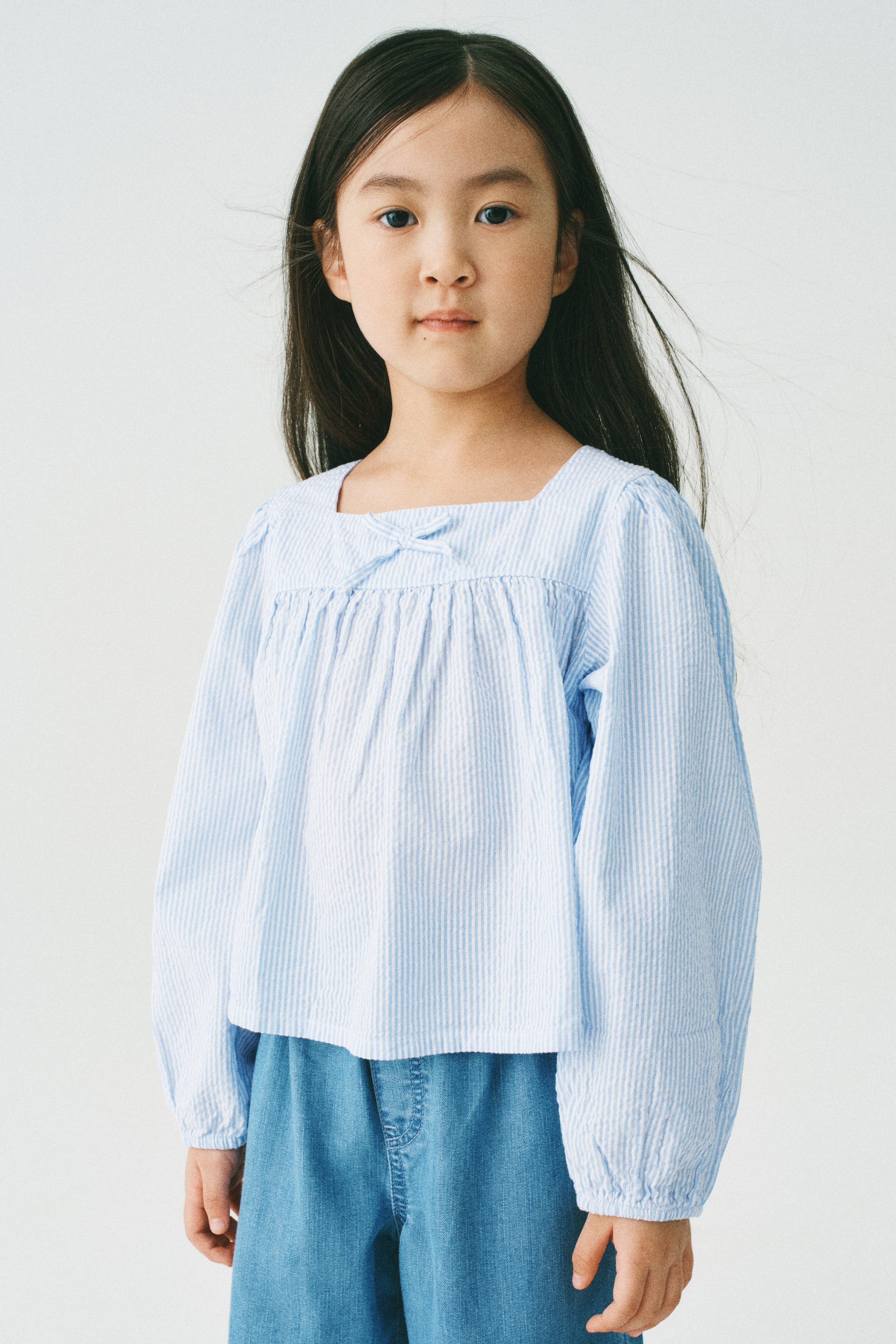 拡大画像を表示: ライトブルーとホワイトのストライプ柄シアサッカーブラウスは、スクエアネックラインに小さなリボンディテールとネックライン下のギャザーが特徴です。長いギャザー入りの袖と、ゆったりとしたクロップドフィットが前向きにデザインされています。