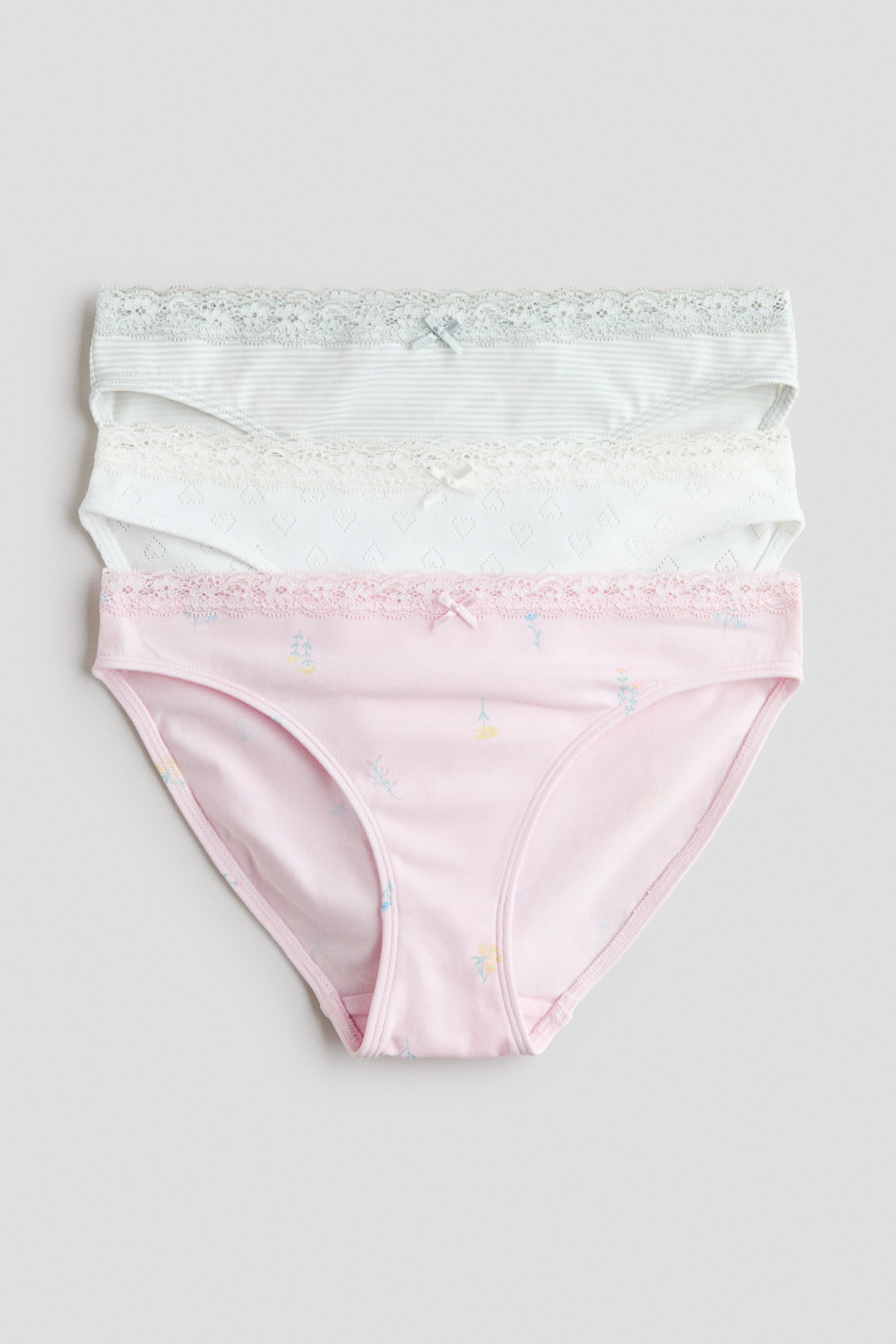 Agrandir l'image: Trois paires de culottes sont empilées face à vous. Chaque culotte est confectionnée en jersey de coton doux, ornée d'un petit nœud en satin décoratif à l'avant et d'une taille élastiquée bordée de dentelle. La culotte du haut présente des rayures vert clair, celle du milieu arbore un motif texturé blanc, et celle du bas affiche un imprimé floral rose.