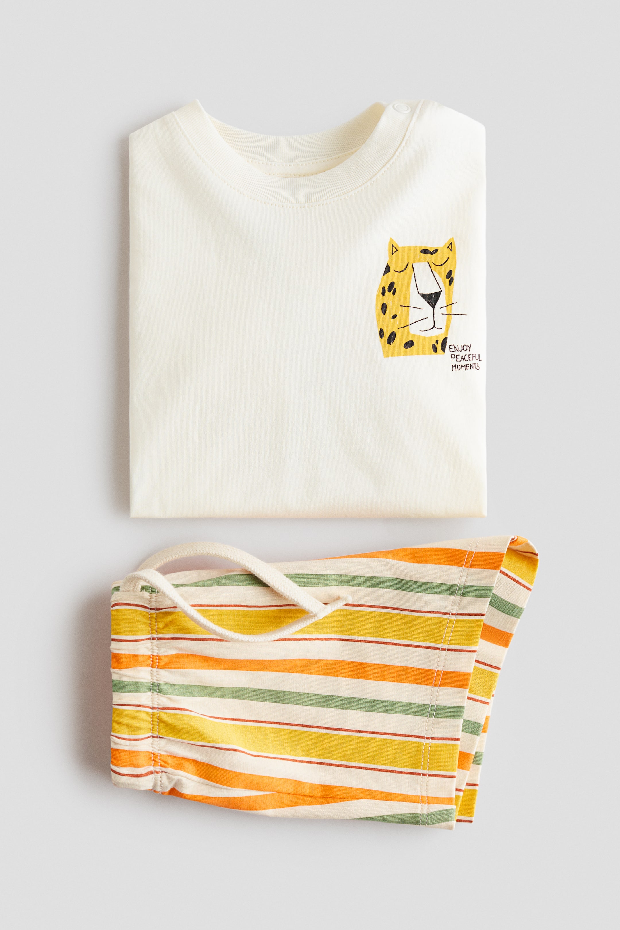 View larger image: A 2-piece cotton set featuring a folded, cream-coloured T-shirt with a round neck, shoulder snaps and a yellow-and-black leopard graphic on the front right chest. The set also includes folded, striped shorts with an elasticated drawstring waist and cream-coloured drawstrings, displaying horizontal stripes in cream, orange, yellow and green.