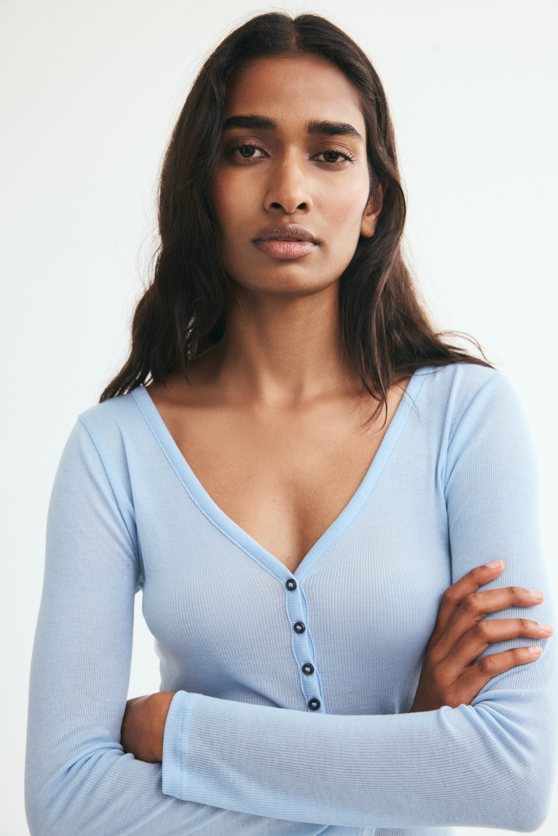 View larger image: A light blue, narrow-ribbed top featuring a low-cut V-neck and a button placket with dark buttons is being worn by the model. The model is posing with crossed arms, highlighting the fitted long sleeves and fine ribbed texture of the garment.
