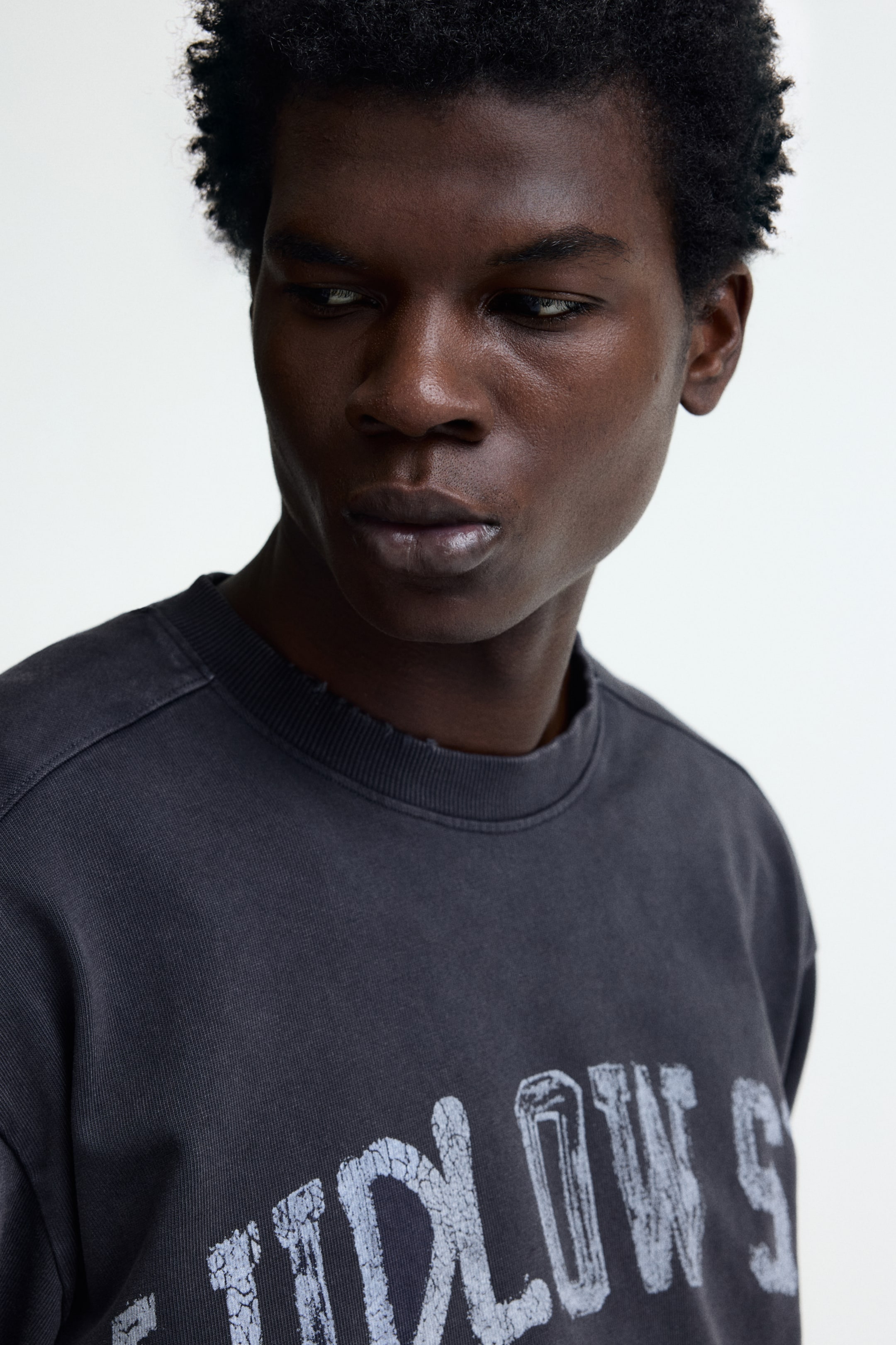 View larger image: A dark gray crew-neck sweatshirt is being shown from an upper chest and shoulder perspective, angled slightly to the viewer's right. It features a distressed ribbed neckline and a washed finish, with faded white lettering partially spelling "LLOW S" on the front.