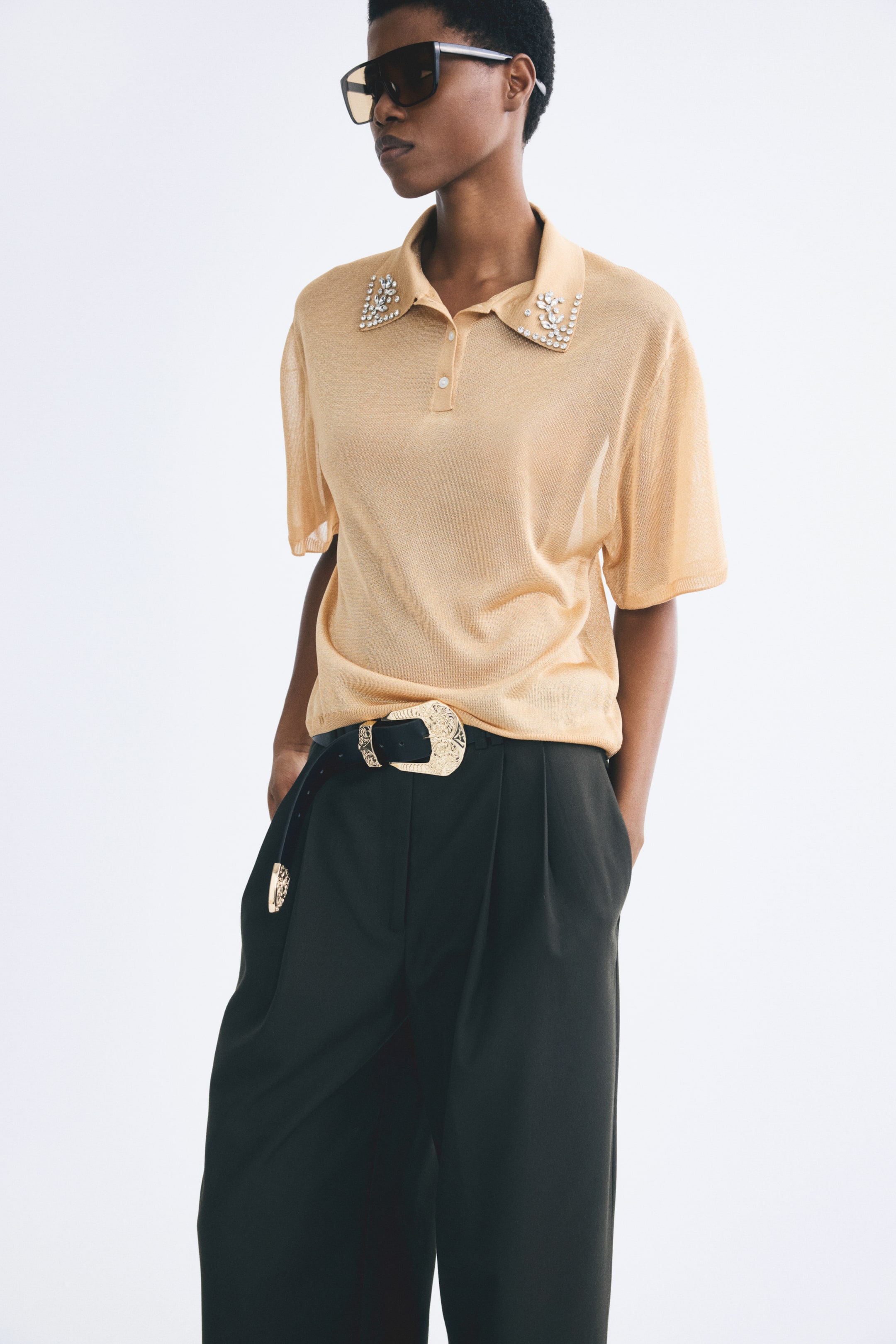 View larger image: A light beige, fine-knit polo shirt is being worn by the model, featuring a collar detailed with sparkling rhinestone embellishments and a button placket. The loose-fit shirt has dropped shoulders and short sleeves, and is tucked into dark trousers styled with a decorative gold-toned belt.