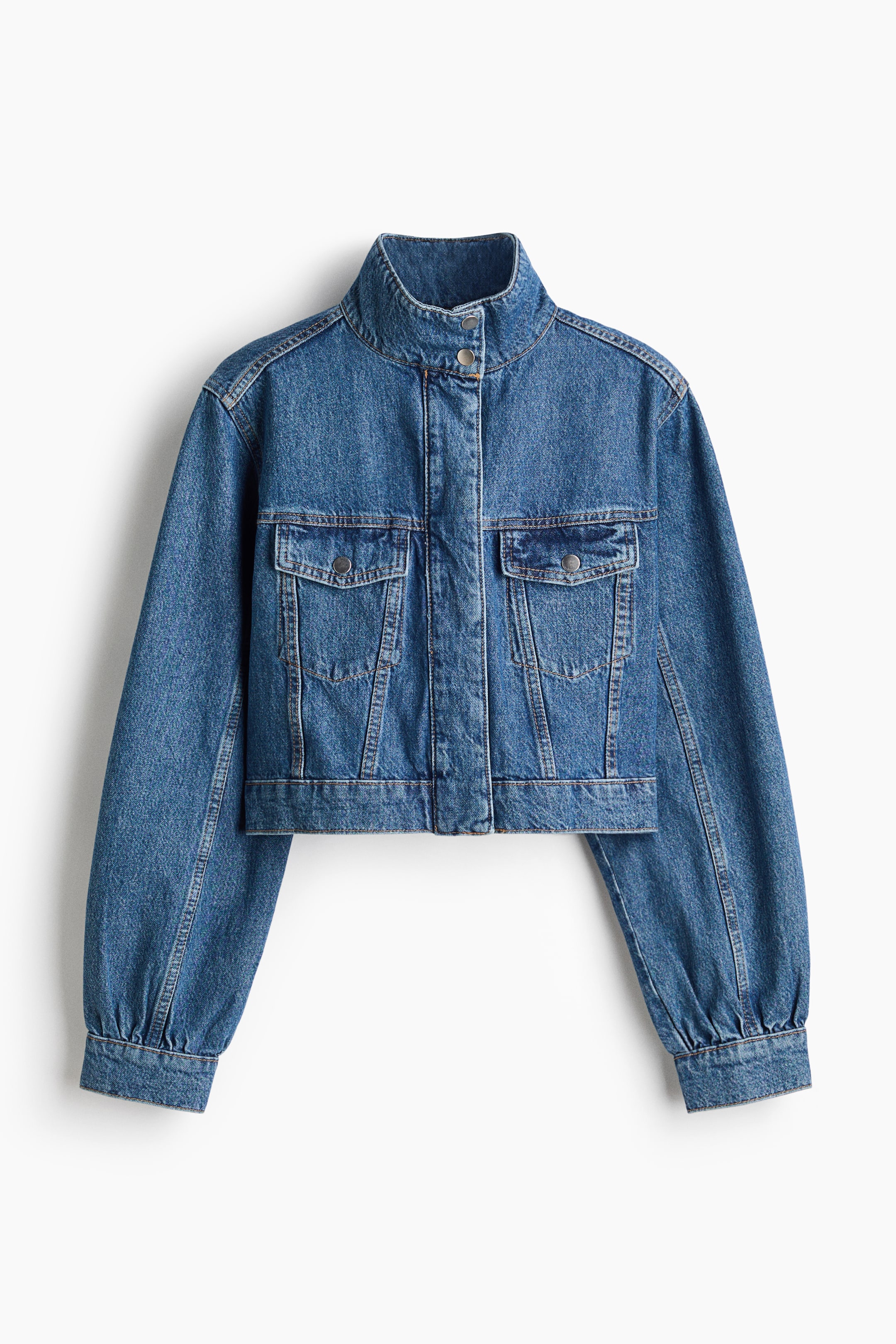 View larger image: A medium blue denim jacket is showcased from the front, highlighting a stand-up collar and a full snap-button placket. It features two chest flap pockets, voluminous long sleeves that gather into fitted cuffs, and a cropped hem, all accented with visible contrast stitching.