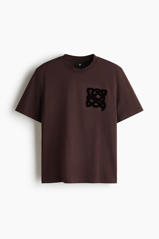 A dark brown cotton-jersey T-shirt is hanging facing forward, featuring a round neckline, short sleeves, and a straight hem. A black, textured interlocking knot graphic details the left side of the chest. - 1