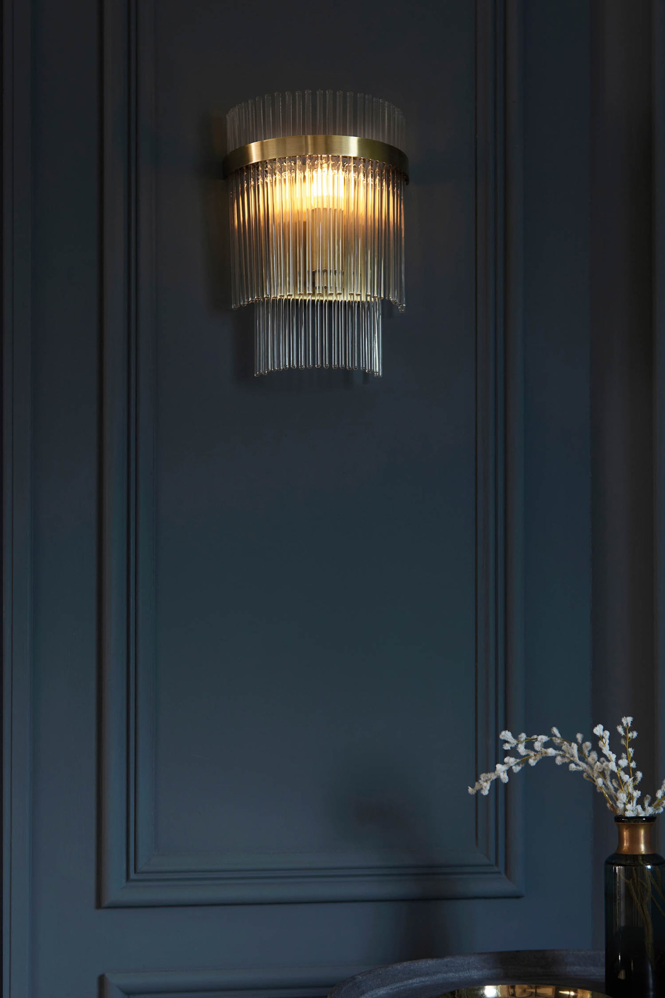 View larger image: A wall sconce featuring a brass-toned metal band supporting multiple clear glass rods arranged in two tiers, with the internal light source brightly illuminating them and casting a warm glow.