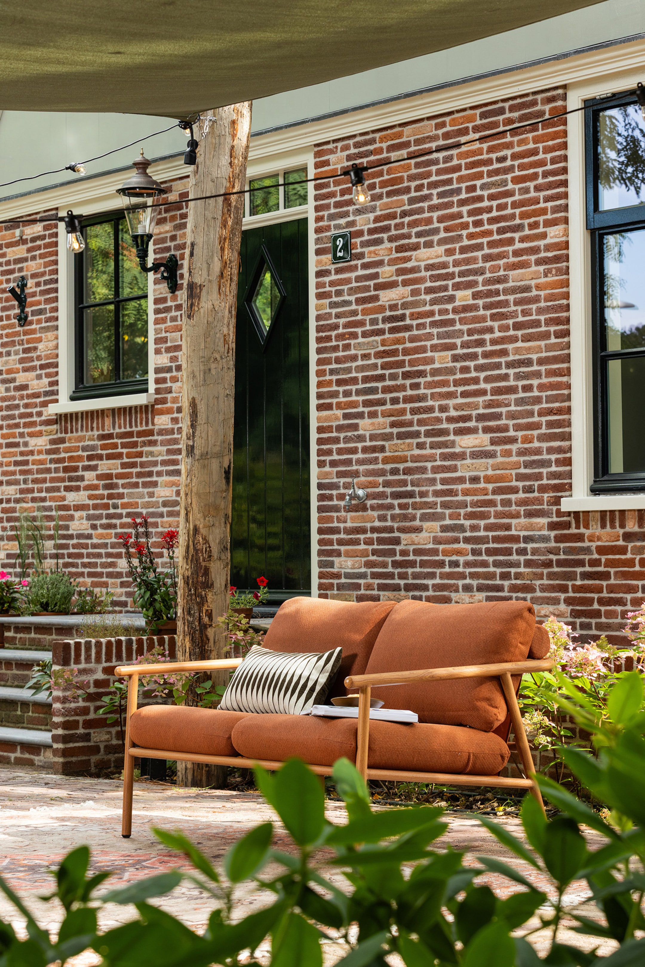 Grotere afbeelding bekijken: Een buitenbank met twee roestoranje kussens en een licht houten frame, lichtjes naar rechts gekanteld. Een zwart-wit geometrisch kussen rust op de linkerzitplaats, en een boek ligt op de rechterzitplaats.