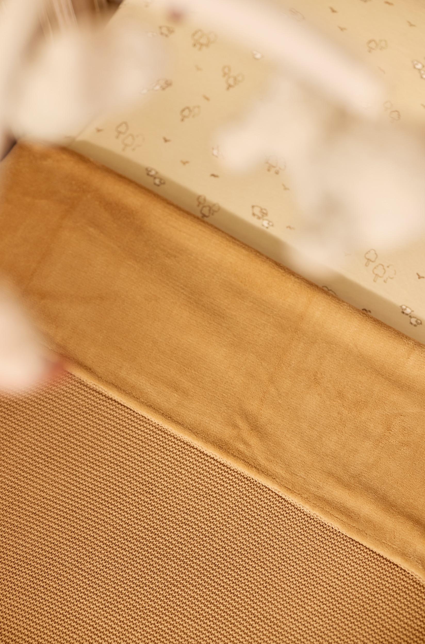 Eine beige Strickdecke liegt flach und ist zu einer sauberen rechteckigen Form gefaltet. Die Decke verfÃ¼gt Ã¼ber ein feines, strukturiertes Strickmuster und zeigt unten links ein kleines quadratisches Etikett mit einem stilisierten âJâ.