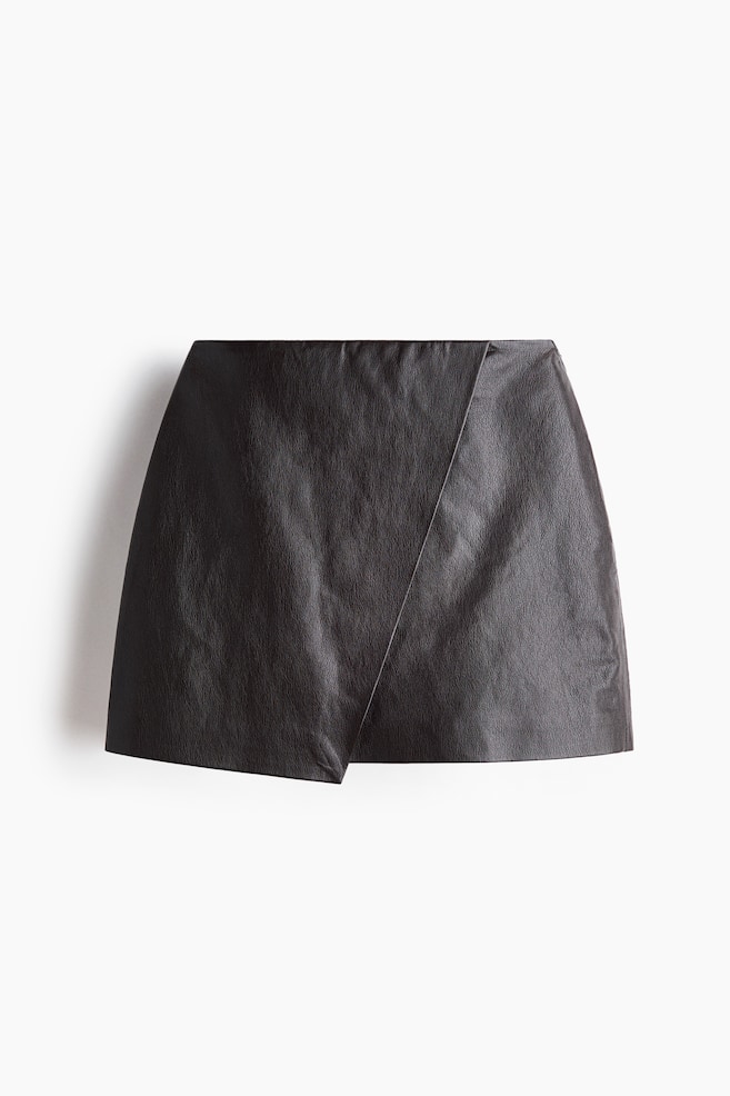 A black faux leather mini skirt is showcased with an asymmetrical wrap design, highlighted by a diagonal seam across the front panel. - 1