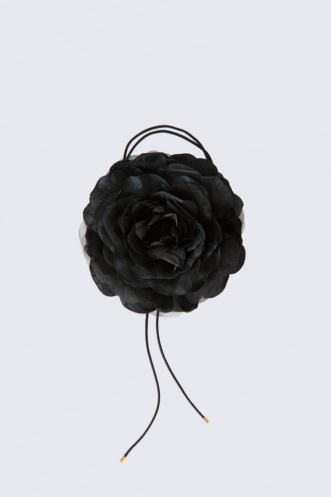 A black fabric flower accessory is shown in a front-facing view, featuring layered, satin-like petals arranged in a rosette shape. Thin black cords form a double loop at the top, while two loose cords with gold-toned tips hang from the bottom. - 1