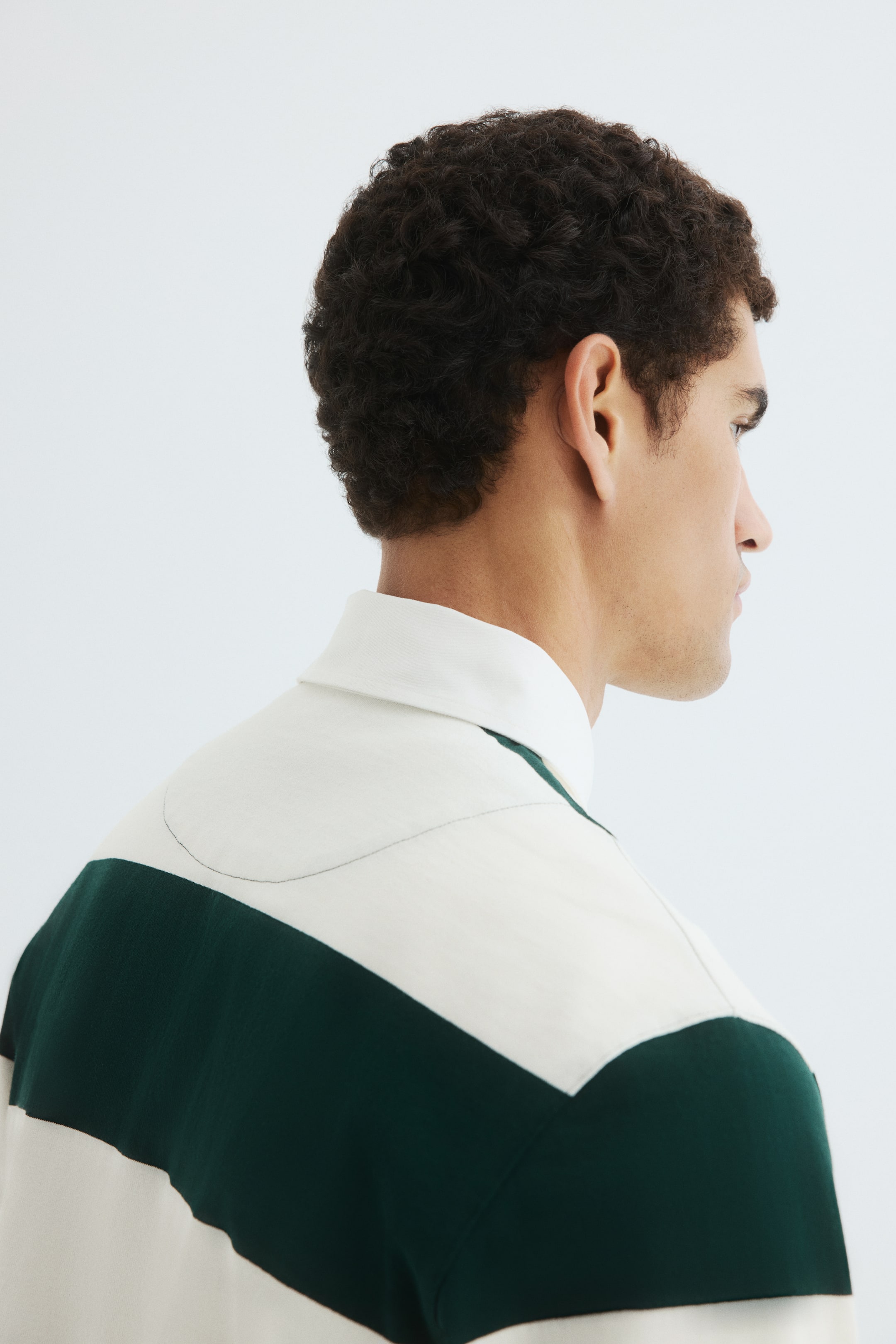 View larger image: A rugby shirt is being viewed from a side and back angle, featuring a white collar. The shirt's upper back is displaying a cream panel with a curved seam, below which a dark green stripe is extending across the shoulder.