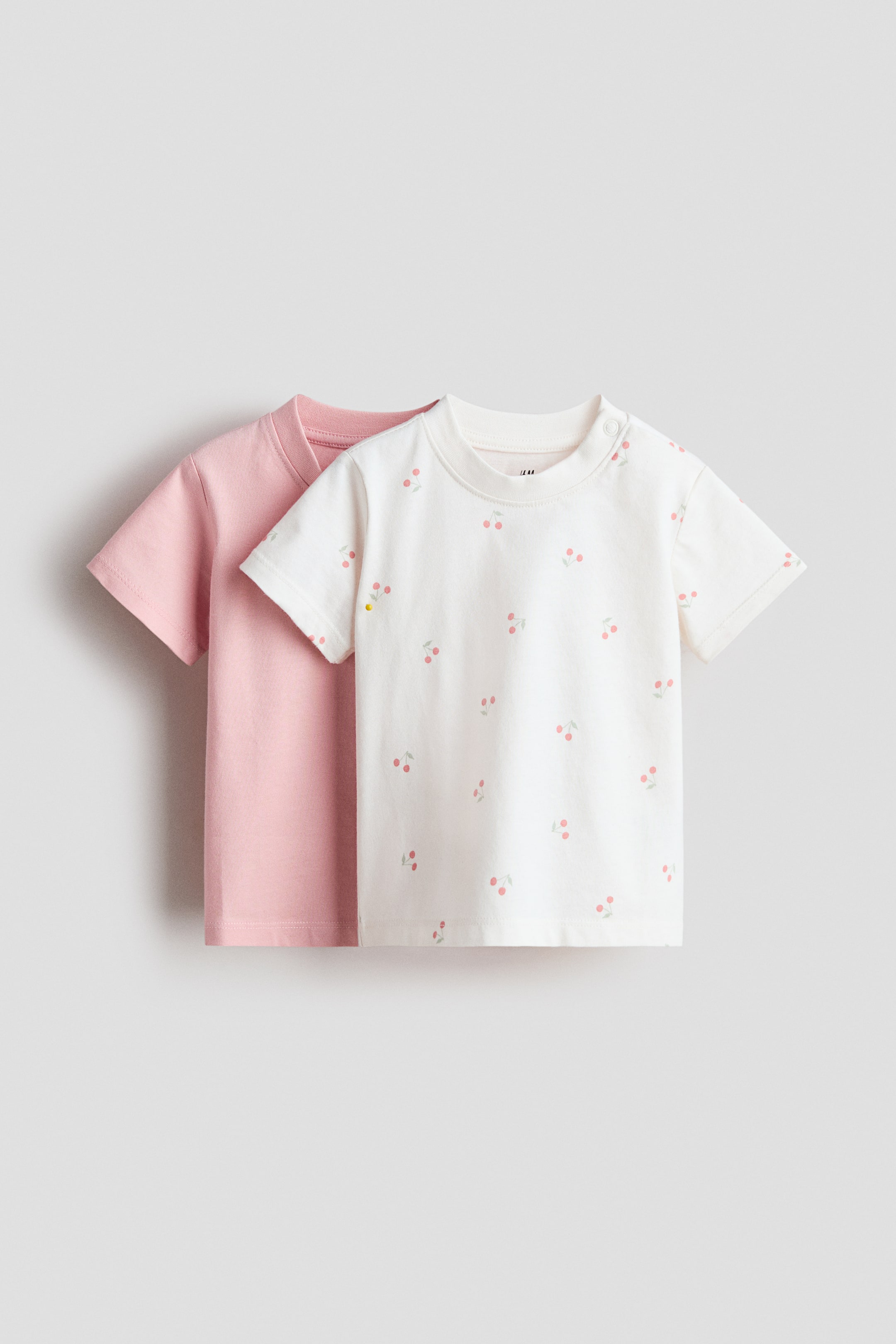 拡大画像を表示: 2枚組のコットンTシャツが展示されています。無地のピンクTシャツがオフホワイトのTシャツの後ろに配置されています。両方のTシャツはショートスリーブで、前面のオフホワイトTシャツには散らばったチェリーパターン、リブ付きクルーネック、左肩に2つのスナップボタンが特徴です。