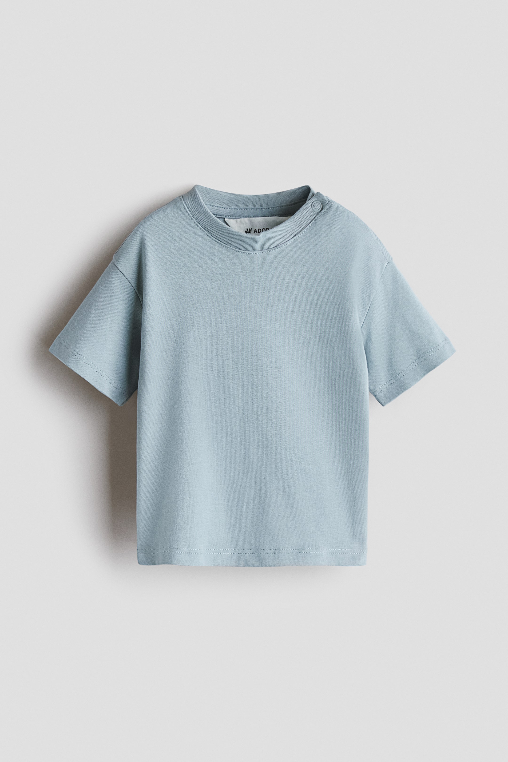 View larger image: A light blue loose-fit T-shirt is displayed from the front, featuring a round, rib-trimmed neckline with a single snap on the left side. It also showcases dropped shoulders and short sleeves.