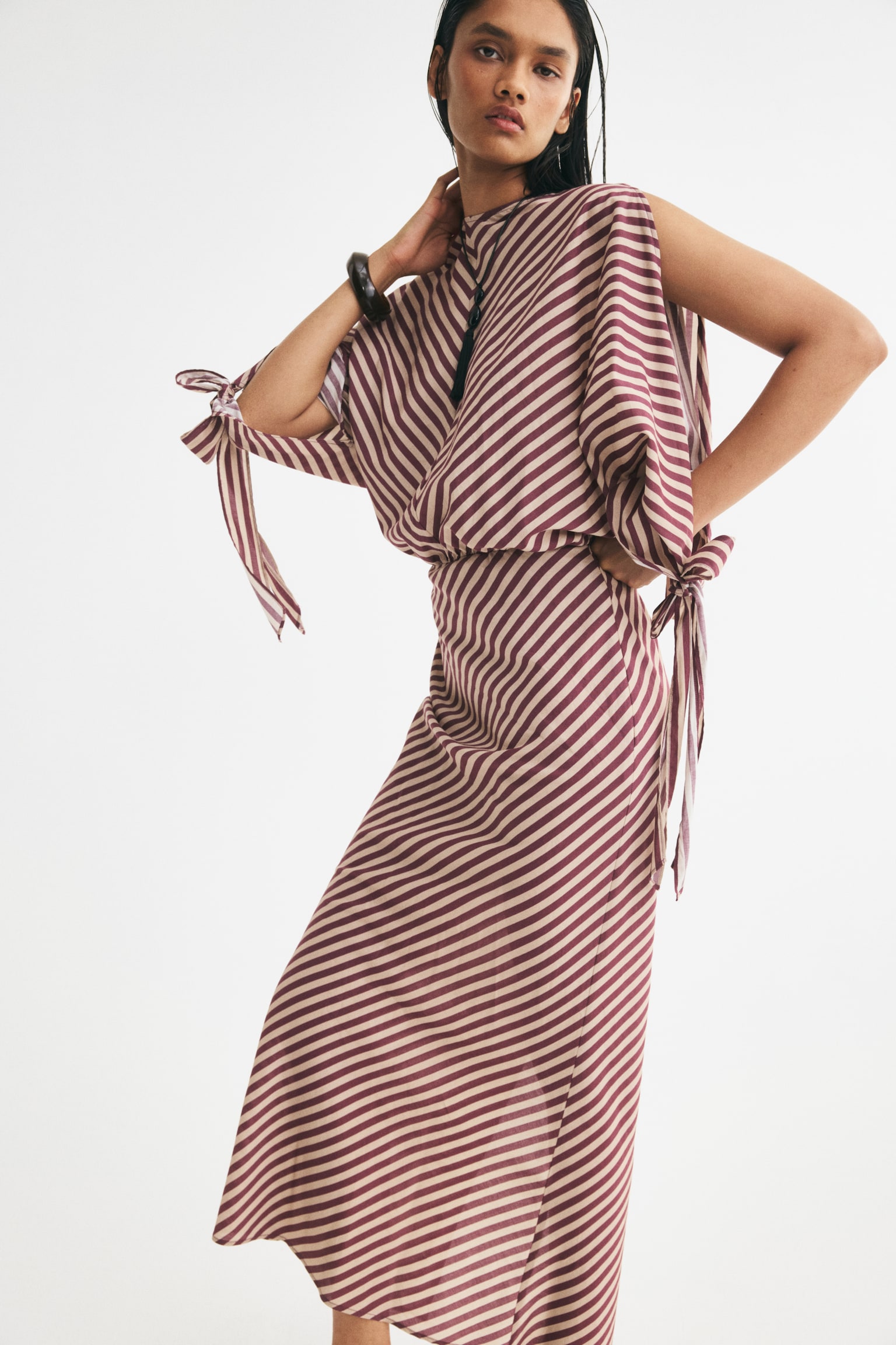 A striped maxi dress showcasing maroon and light pink diagonal stripes, creating a chevron pattern on the V-neck bodice and extending diagonally down the skirt. It is crafted with long, loose sleeves, each adorned with a tie-cuff, and features an elasticated waist. - 1