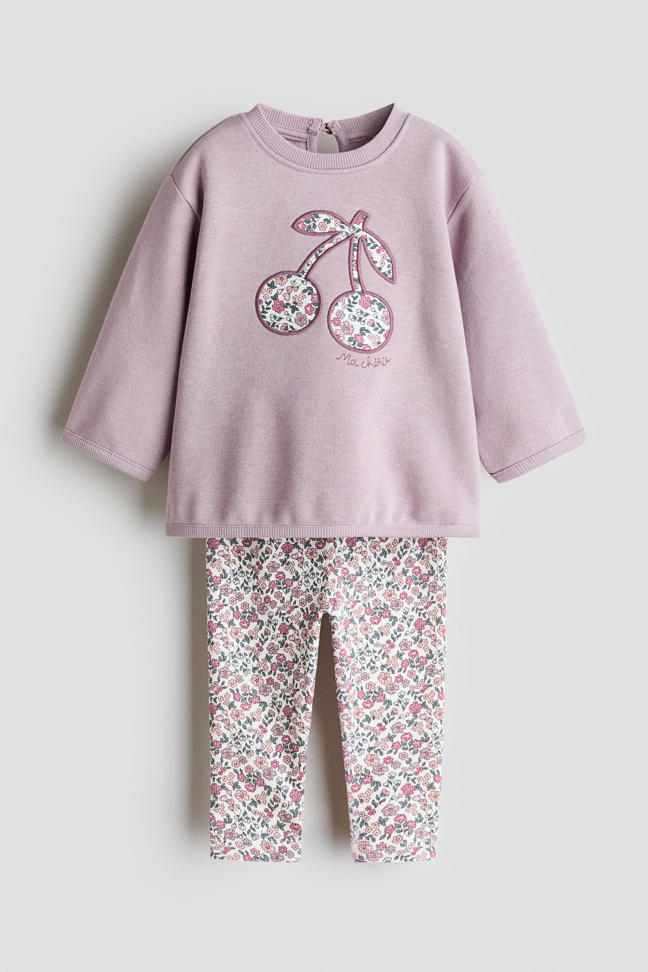 View larger image: The two-piece children's clothing set features a lilac sweatshirt and matching floral leggings, presented facing front. The lilac sweatshirt showcases a crew neck, ribbed cuffs and hem, and a button closure at the back of the neck, with a large cherry appliqué decorating its front. The cherry appliqué displays a small floral pattern on its fruit and leaves, and "Ma chérie" is embroidered below it; the matching leggings exhibit an allover small floral print in pink, green, and white on a light background.