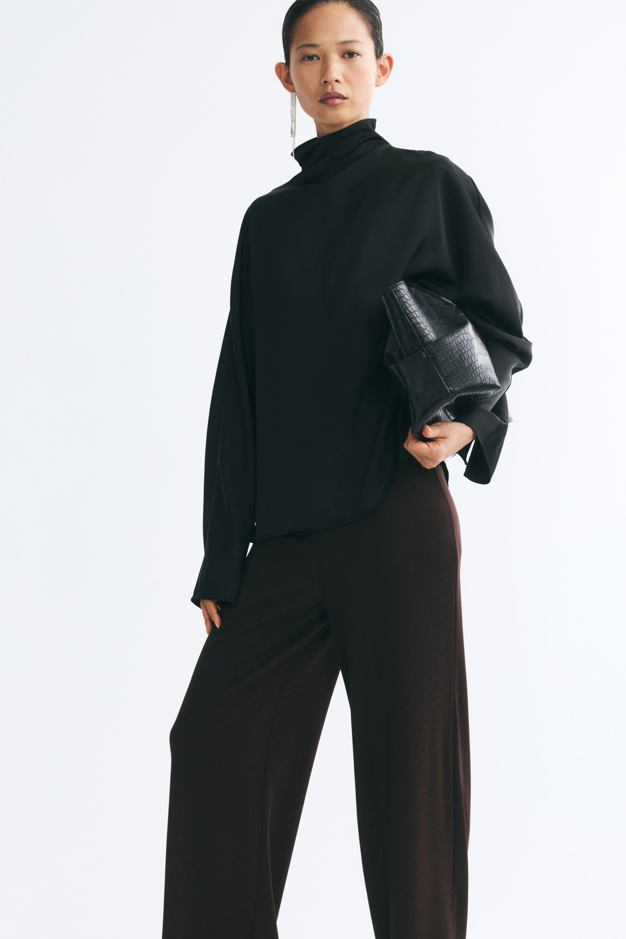 Agrandir l'image: Un pantalon ample à jambes larges en maille marron foncé est porté par le mannequin, avec une coupe décontractée. Le pantalon est associé à un haut noir à col montant et à une pochette noire texturée.