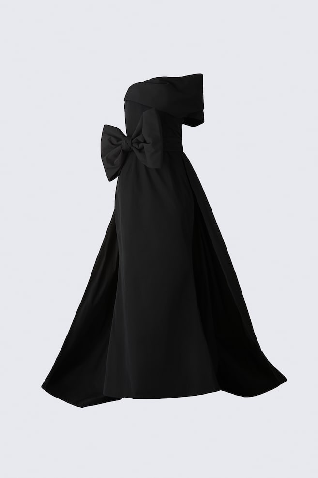 Un abito nero lungo fino a terra con scollo a fascia è mostrato da un'angolazione laterale tre quarti, mettendo in risalto la sua silhouette strutturata. L'abito presenta un pannello a manica drappeggiato con spalle scoperte, un grande fiocco coordinato sul fianco in vita e una gonna ampia svasata con un drammatico strascico. - 1