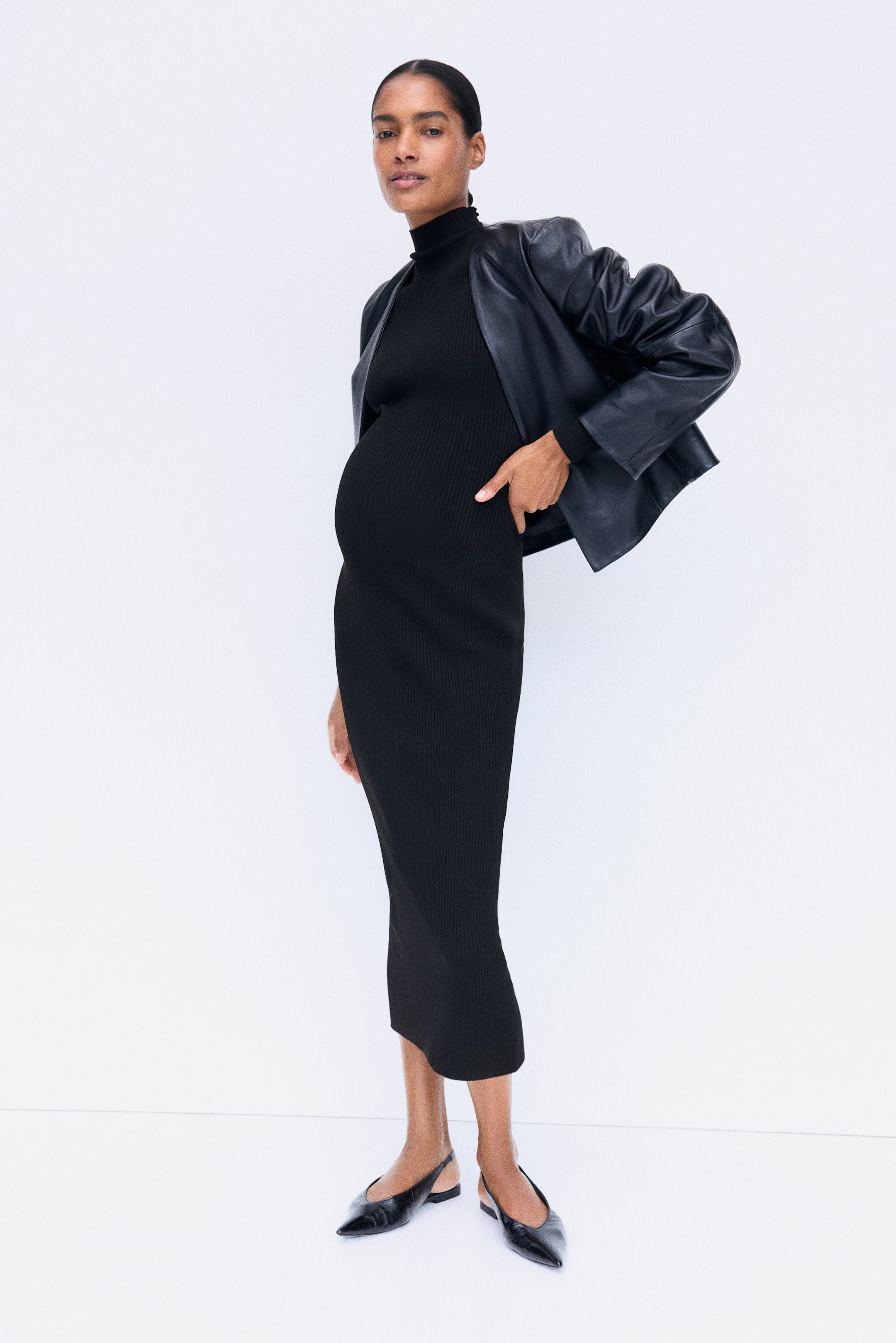 Ampliar la imagen: La modelo lleva puesto un vestido largo negro ajustado de punto canalé, con cuello alto y mangas largas. El vestido se complementa con una chamarra de piel negra echada sobre los hombros y zapatos planos negros tipo slingback.