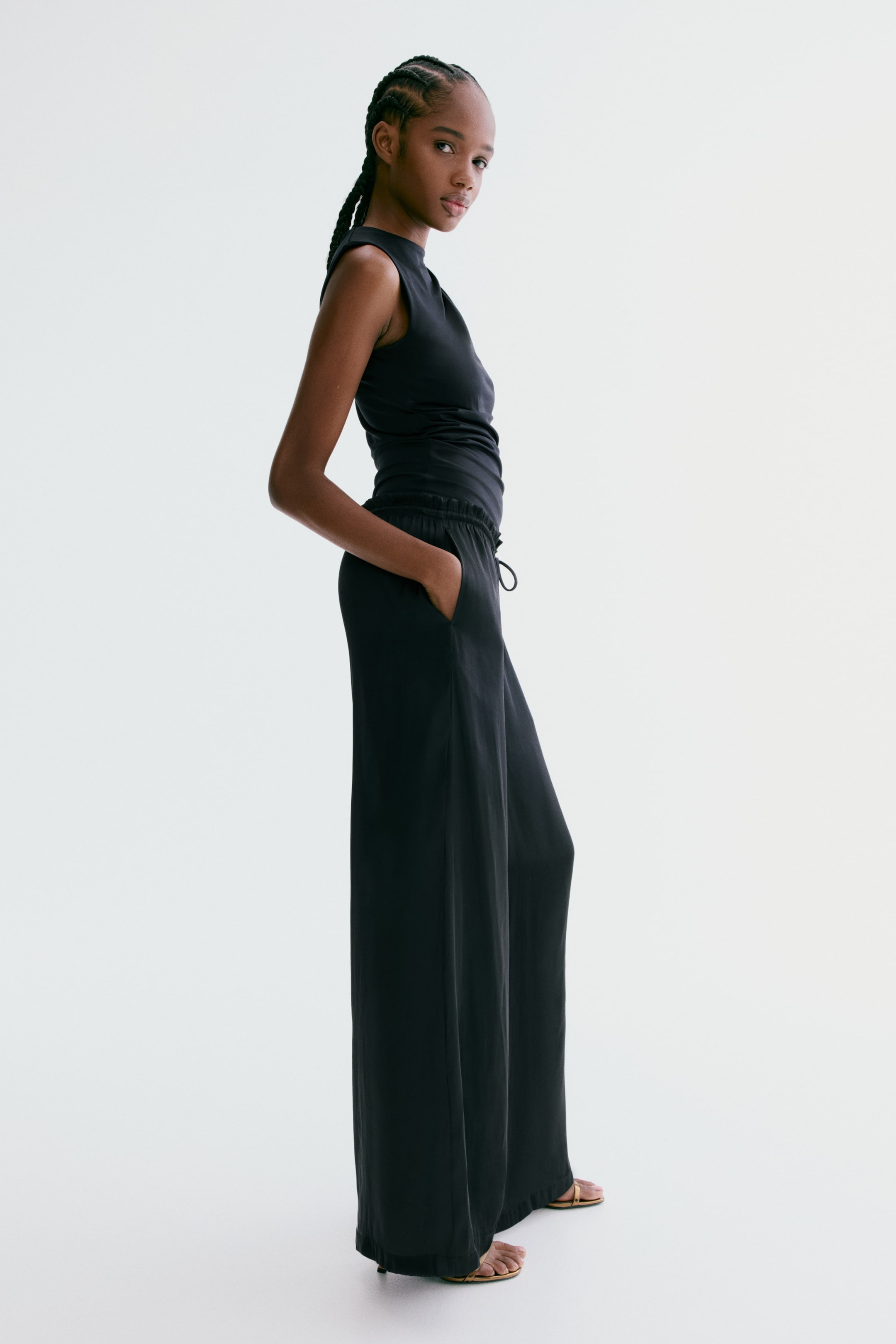 הצגת תמונה גדולה יותר: המכנסיים הרחבים עשויים סאטן שחור רך, נלבשים על ידי הדוגמנית, וכוללים מותן עם שרוך, כיסים צדדיים דיסקרטיים ורגליים רחבות הנופלות מעל הרגל עם אפקט ערימה. הדוגמנית עומדת, עם יד אחת בכיס, ולובשת גם חולצה שחורה ללא שרוולים וסנדלים שטוחים.