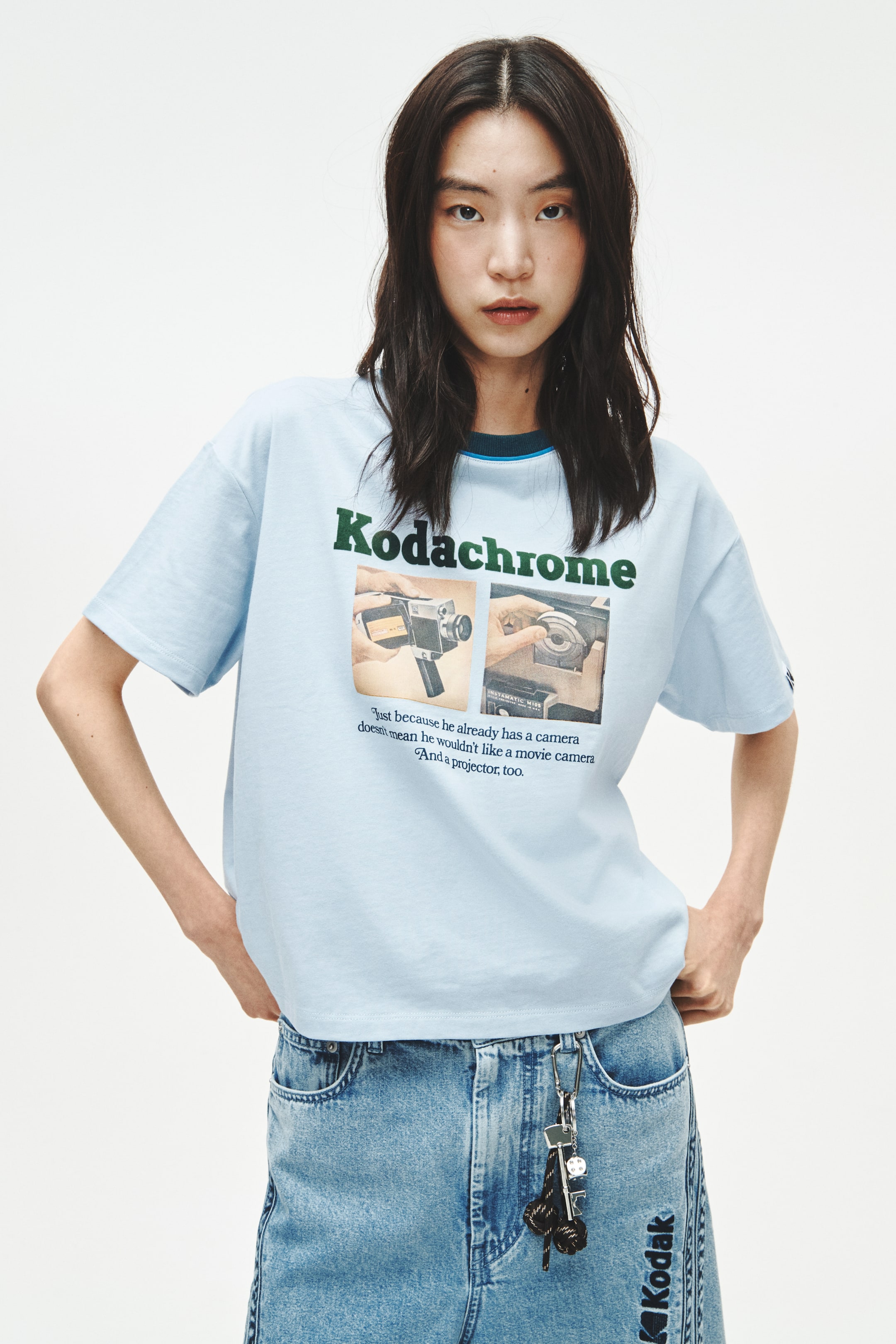 View larger image: A light blue t-shirt features a crew neck with a dark blue ribbed collar, accented by a light blue stripe. The front showcases a large graphic print, including "Kodachrome" in dark green, alongside two square images of hands operating vintage camera equipment, with a quote below them.