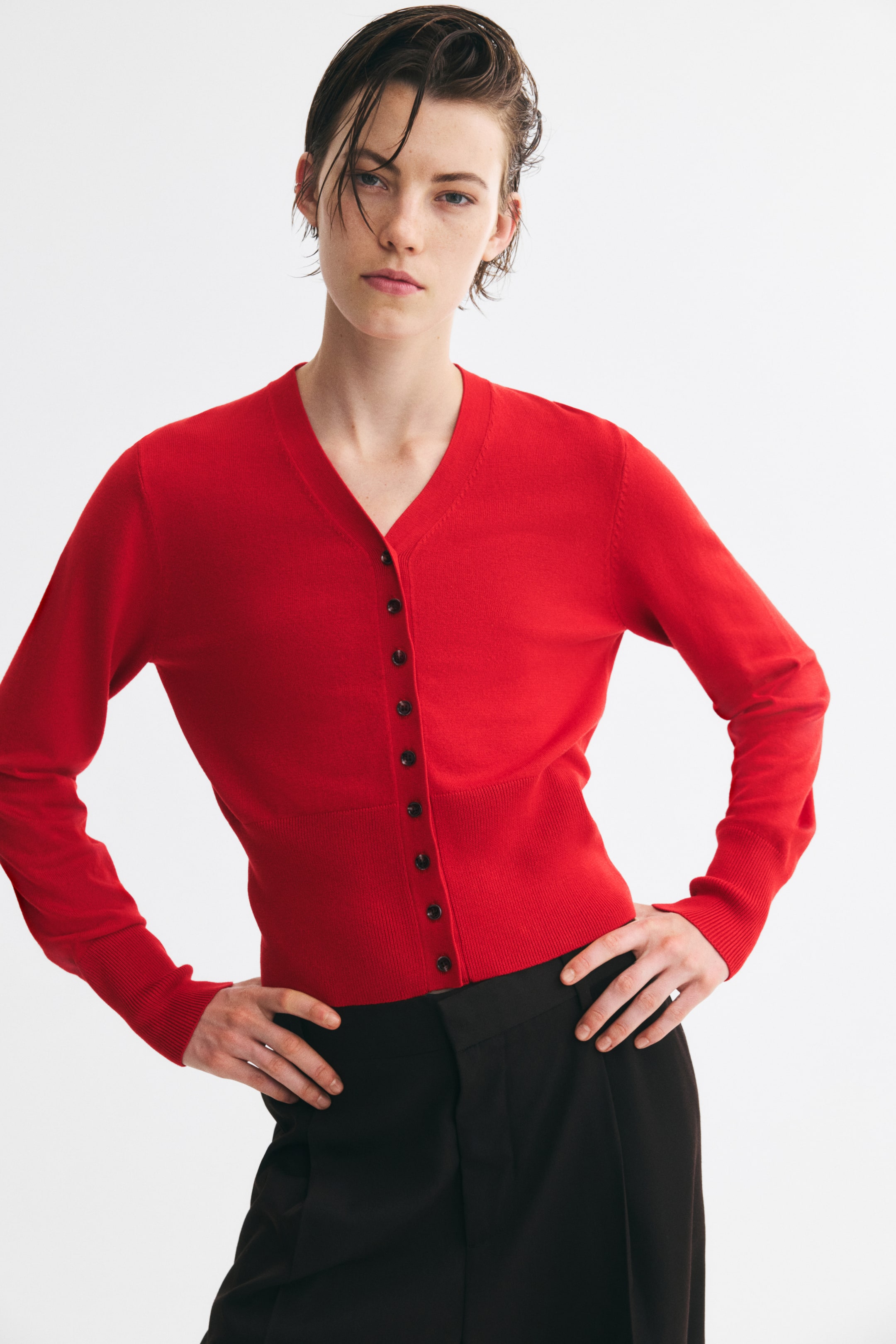 Agrandir l'image: Un cardigan court et ajusté en maille fine, présenté de face, avec un col en V et de petits boutons noirs sur le devant. Le cardigan est porté boutonné, mettant en valeur des manches longues avec de larges poignets côtelés et un large bord-côte à la base.
