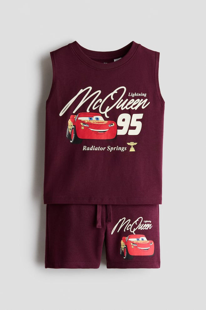 Facing the front, this kids' maroon two-piece clothing set is featuring a sleeveless crewneck top and matching drawstring shorts. The top is displaying a large graphic print of Lightning McQueen with the text "McQueen" and "95", while the shorts are showing a smaller matching graphic on the left leg. - 1