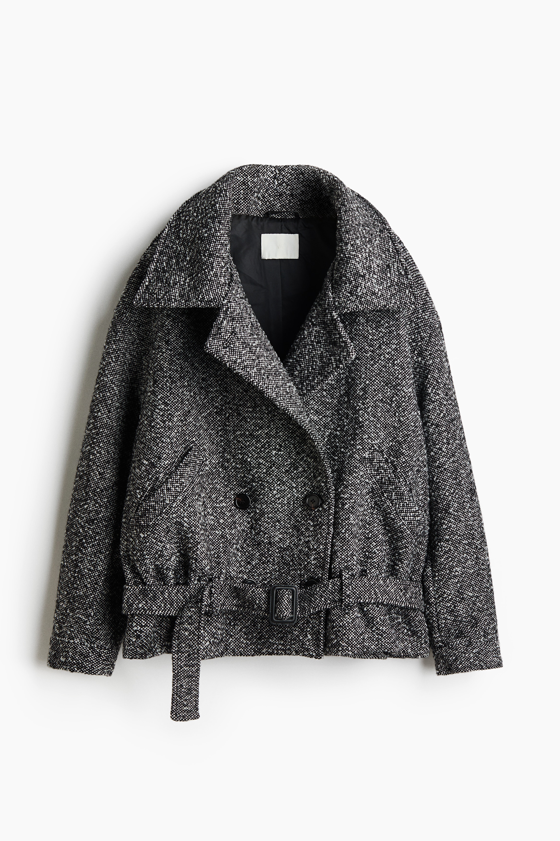 An oversized, double-breasted jacket in a textured weave is worn by the model, with their hands tucked into the front welt pockets. The jacket features a high collar, dropped shoulders, button fastenings, and an adjustable belt with a buckle near the hem. It is styled with wide-leg trousers and dark shoes.