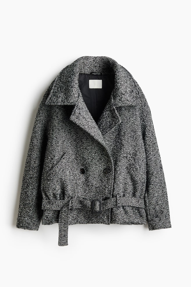 Eine schwarz-weiÃ melierte Tweedjacke ist von vorne zu sehen. Sie verfÃ¼gt Ã¼ber breite fallende Revers und eine zweireihige Vorderseite mit dunklen KnÃ¶pfen. Die Jacke zeigt einen passenden StoffgÃ¼rtel mit schwarzer Schnalle am Saum, schrÃ¤ge Leistentaschen und ein schwarzes Innenfutter. - 1