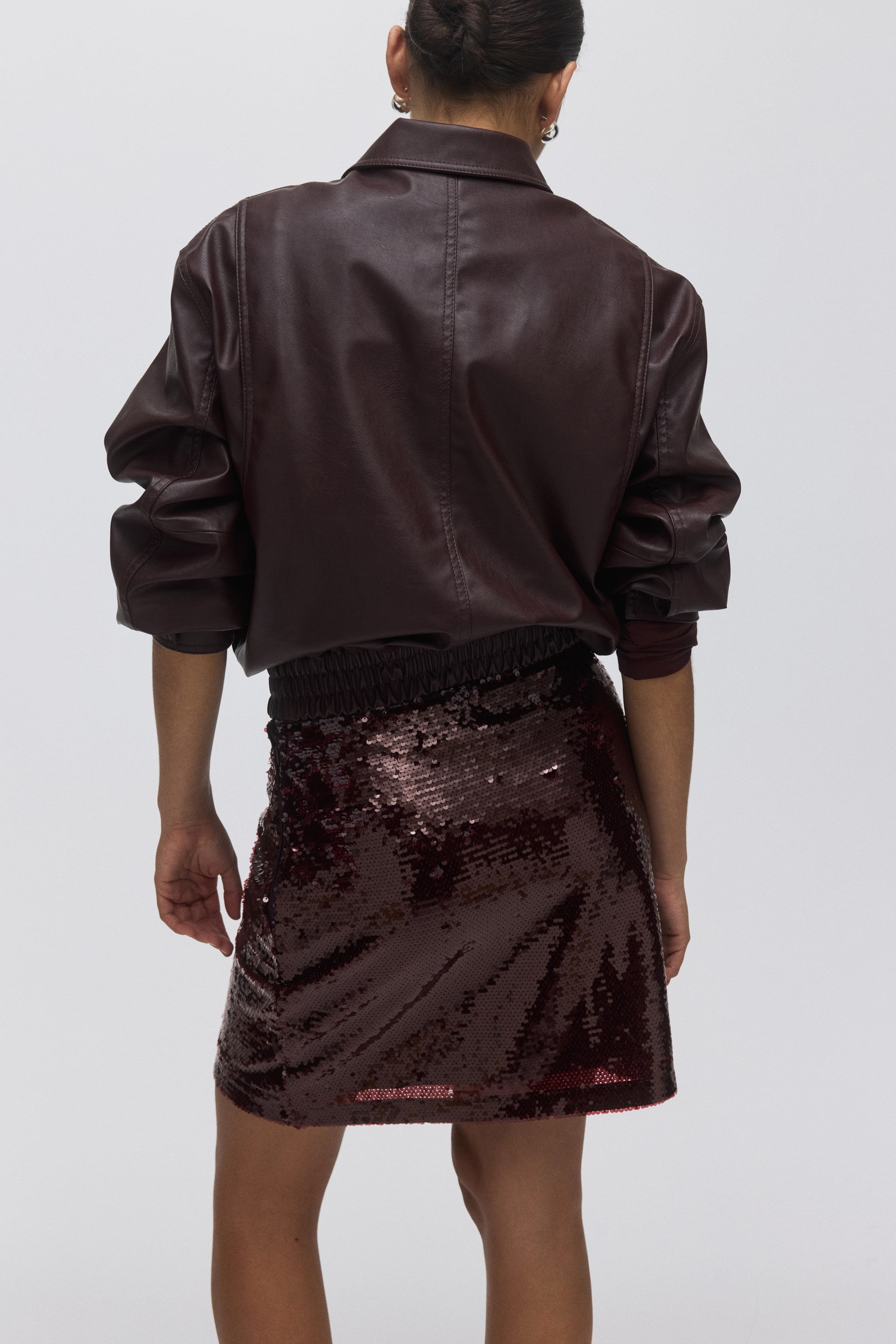 View larger image: A dark reddish-brown faux leather jacket is seen from the back, featuring a collar, gathered sleeves with volume at the elbows, and a gathered elasticated hem. It is paired with a matching sequined mini skirt, which is featuring an elasticated, ruched waistband.