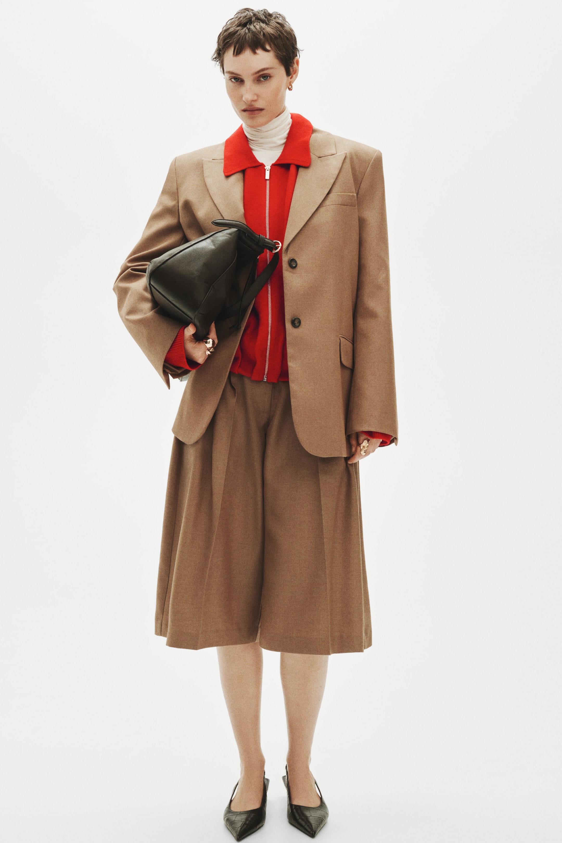Größeres Bild anzeigen: Ein brauner, weit geschnittener Blazer mit Kerbkragen und Zweiknopfverschluss liegt über einem roten Reißverschluss-Sweater und einem weißen Rollkragenpullover und wird mit passenden Culottes mit weitem Bein kombiniert. Eine schwarze Handtasche aus weichem Leder wird unter dem linken Arm getragen, zum Betrachter links geneigt, schwarze spitze Schuhe vervollständigen das Ensemble.