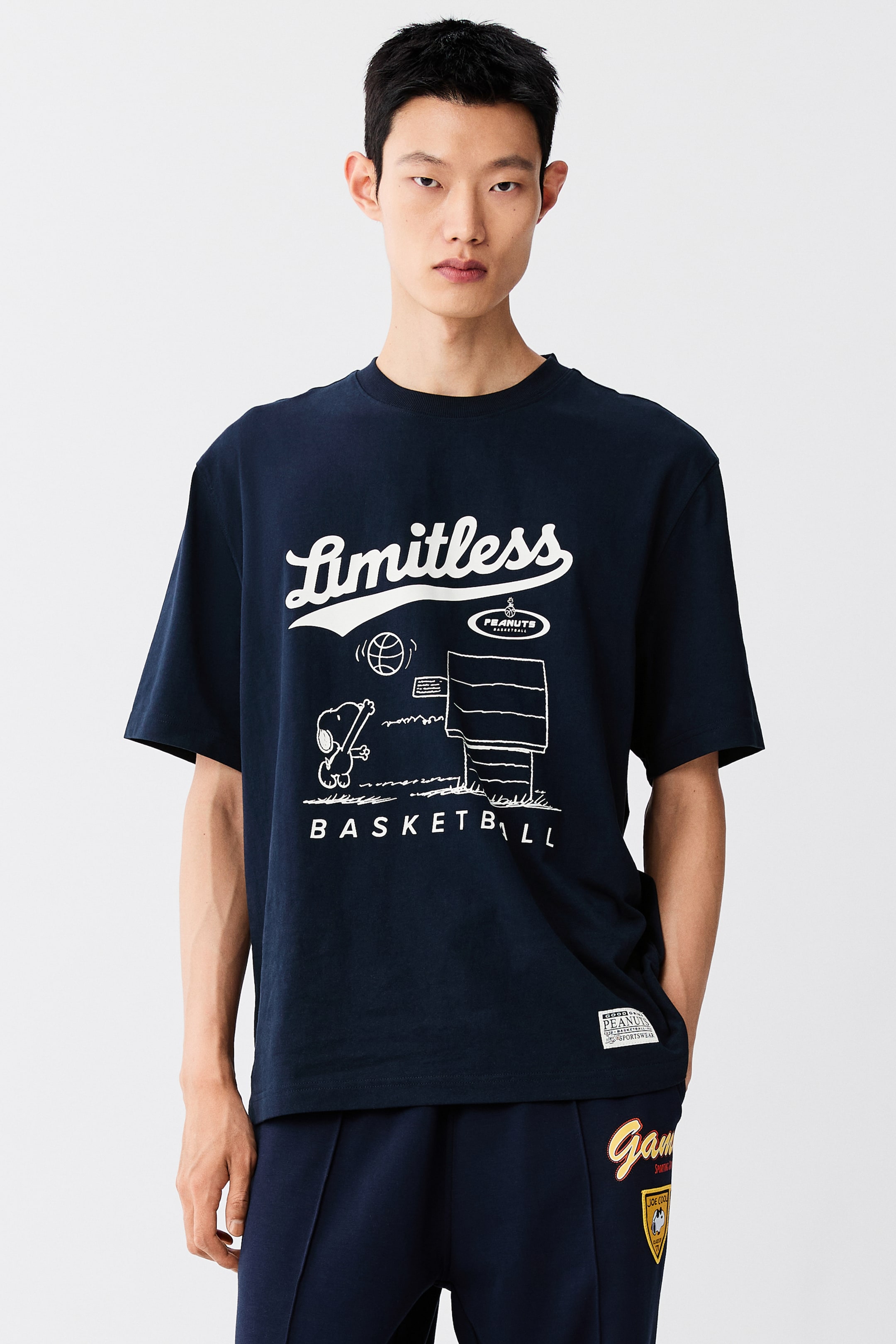 이미지 확대 보기: 모델이 스누피가 농구하는 화이트 그래픽 프린트와 "Limitless" 텍스트가 돋보이는 다크 네이비 블루 티셔츠를 착용하고 있습니다. 루즈핏 티셔츠는 골지 크루넥과 드롭 숄더 디테일이 특징이며, 같은 네이비 블루 스웨트팬츠와 함께 스타일링되었습니다.