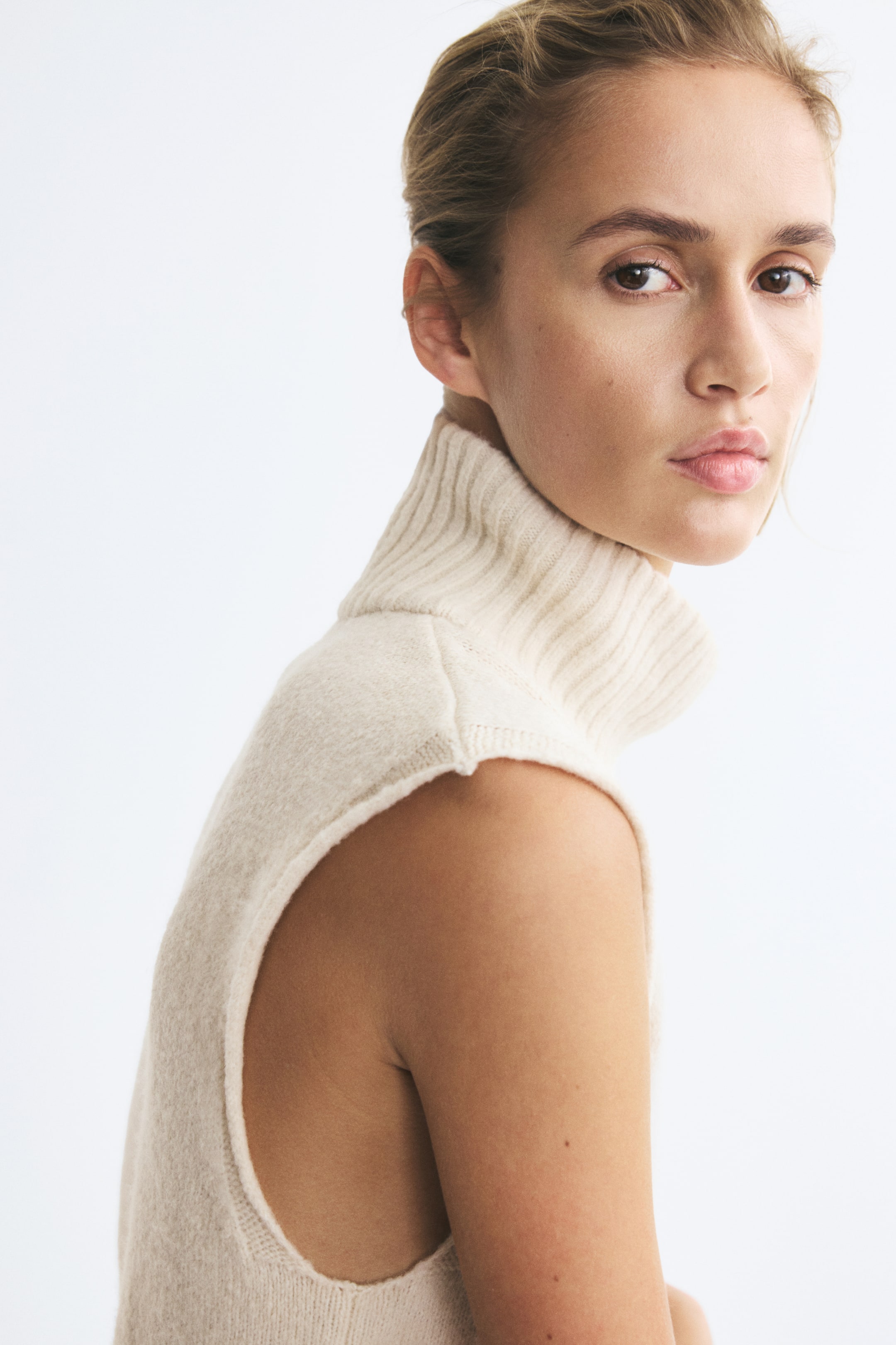 Visualizza immagine più grande: Il modello indossa un morbido gilet in maglia color crema con collo alto a coste e giromanica senza maniche con bordi arrotolati.
