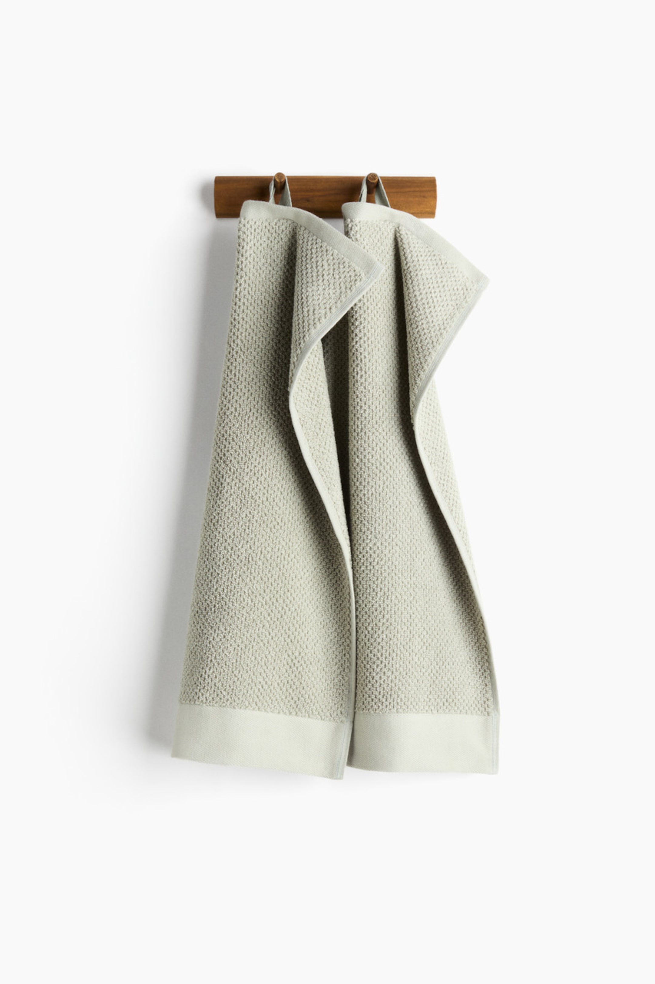 View larger image: Two light grey guest towels hang side-by-side, each showcasing a textured dobby weave pattern. Each towel is adorned with a smooth, flat woven border along the short ends and is suspended by a fabric loop at one short side.
