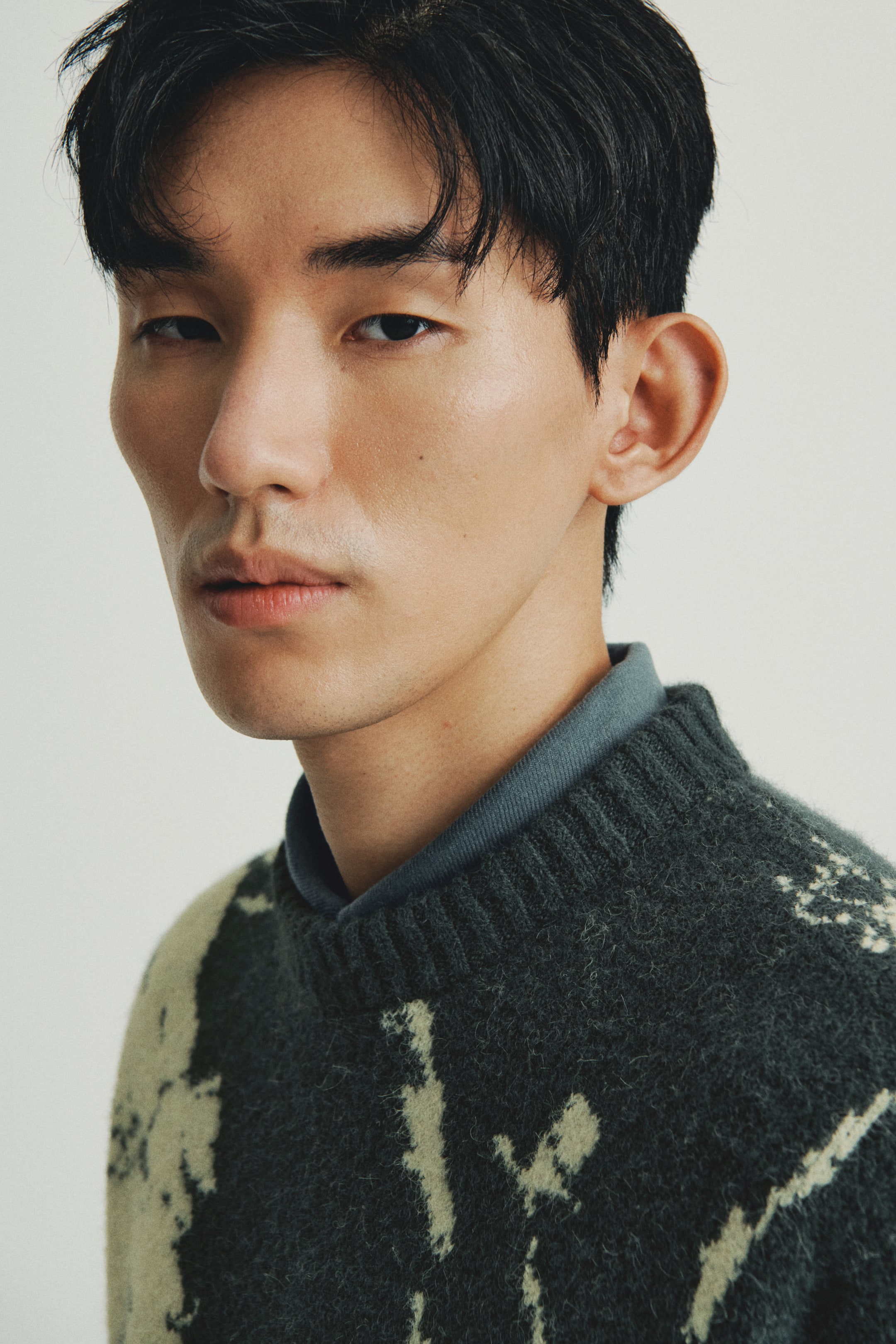 이미지 확대 보기: 짙은 녹색의 보송보송한 크루넥 스웨터가 착용되어 있으며, 리브드 넥라인과 불규칙한 밝은 색조의 추상적인 패턴이 특징입니다. 스웨터 넥라인 아래로 짙은 녹색의 칼라 셔츠가 보입니다.