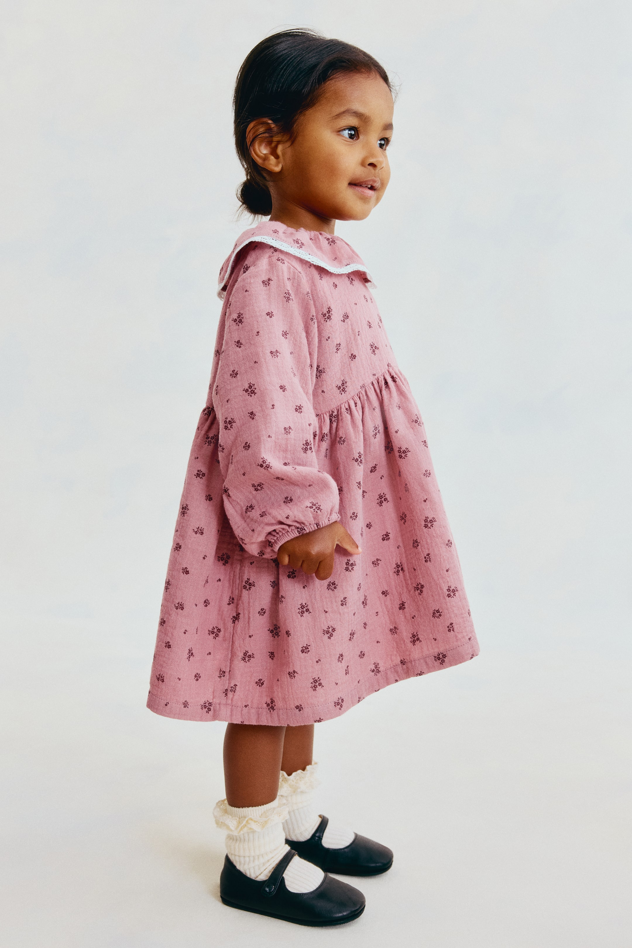 View larger image: A dusty pink cotton dress featuring a dark floral pattern is shown from a side profile facing right, highlighting its flared silhouette. The dress features a round collar with white lace trim, long raglan sleeves with narrow elastic cuffs, and a high gathered waist.